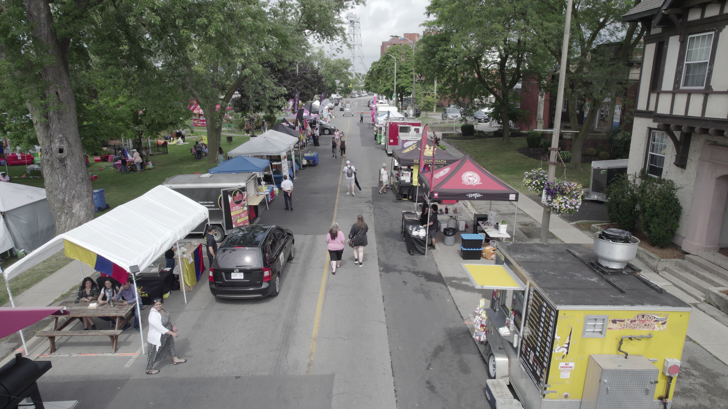 Aerial view of a street fair with various food trucks, tents, and people walking and shopping on a street lined with trees and buildings.