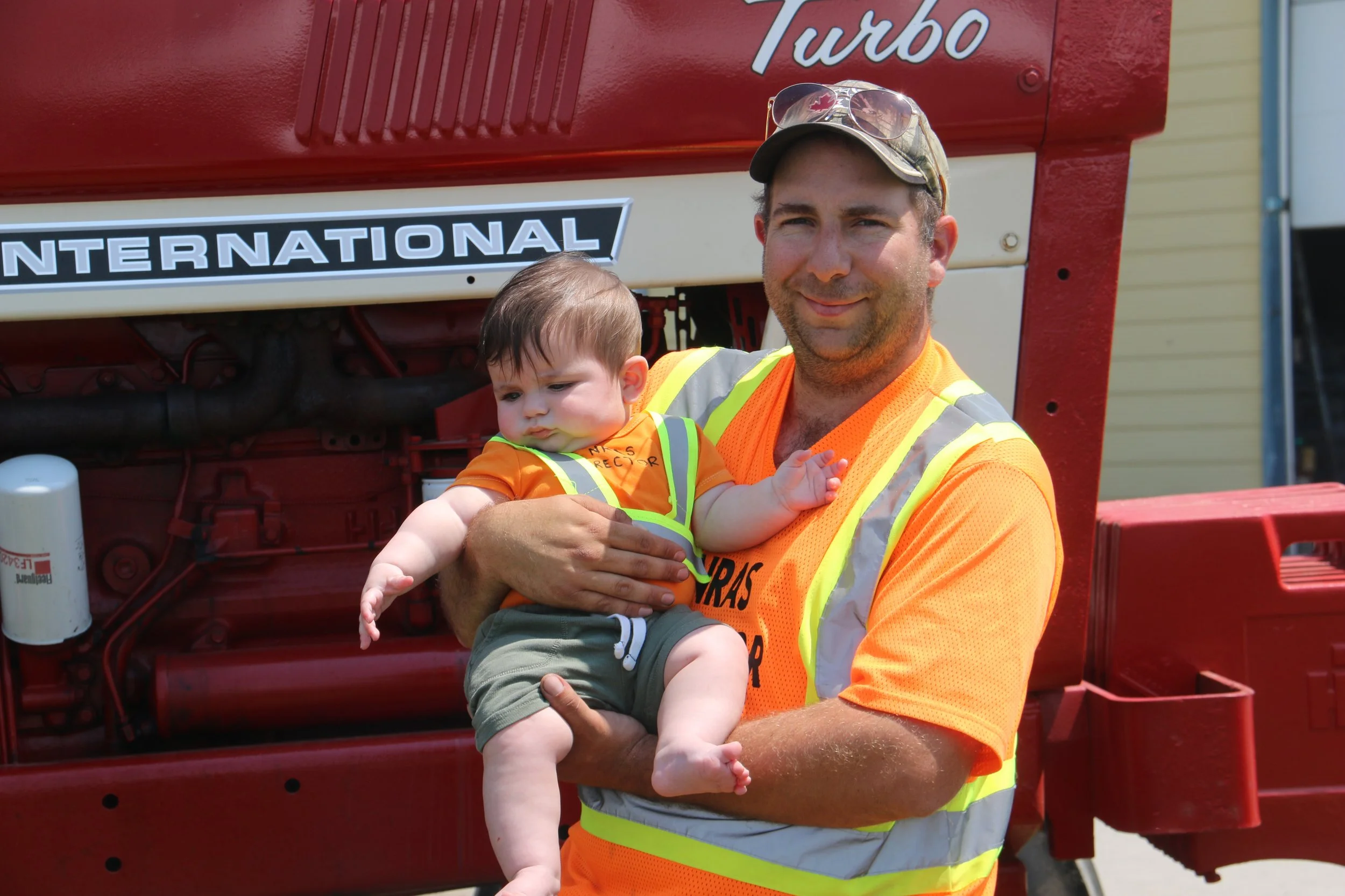 A man in an orange safety vest holding a baby, standing in front of a red fire truck.