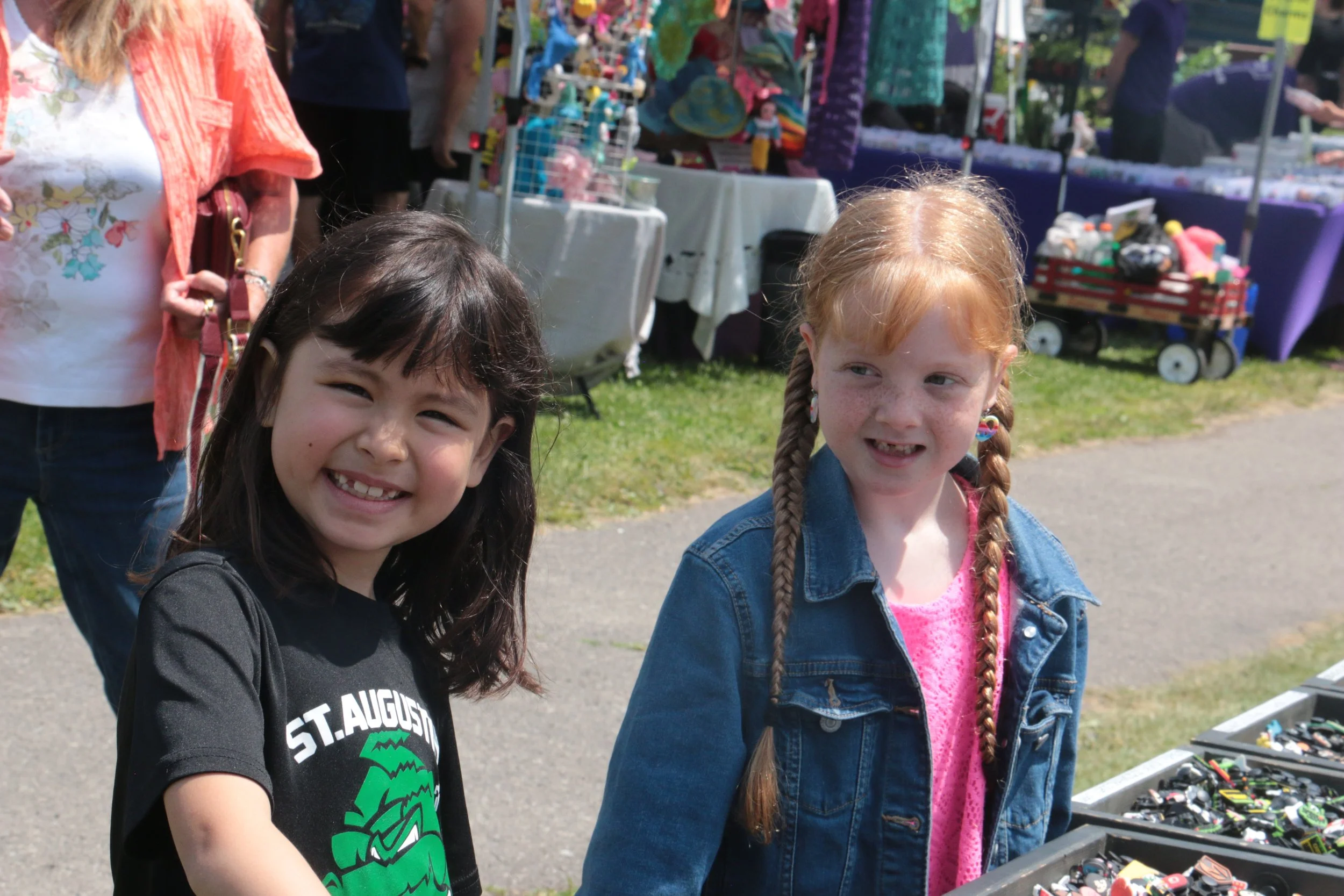 Two young girls smiling at an outdoor market with various items on display, including jewelry and toys, under sunny weather.