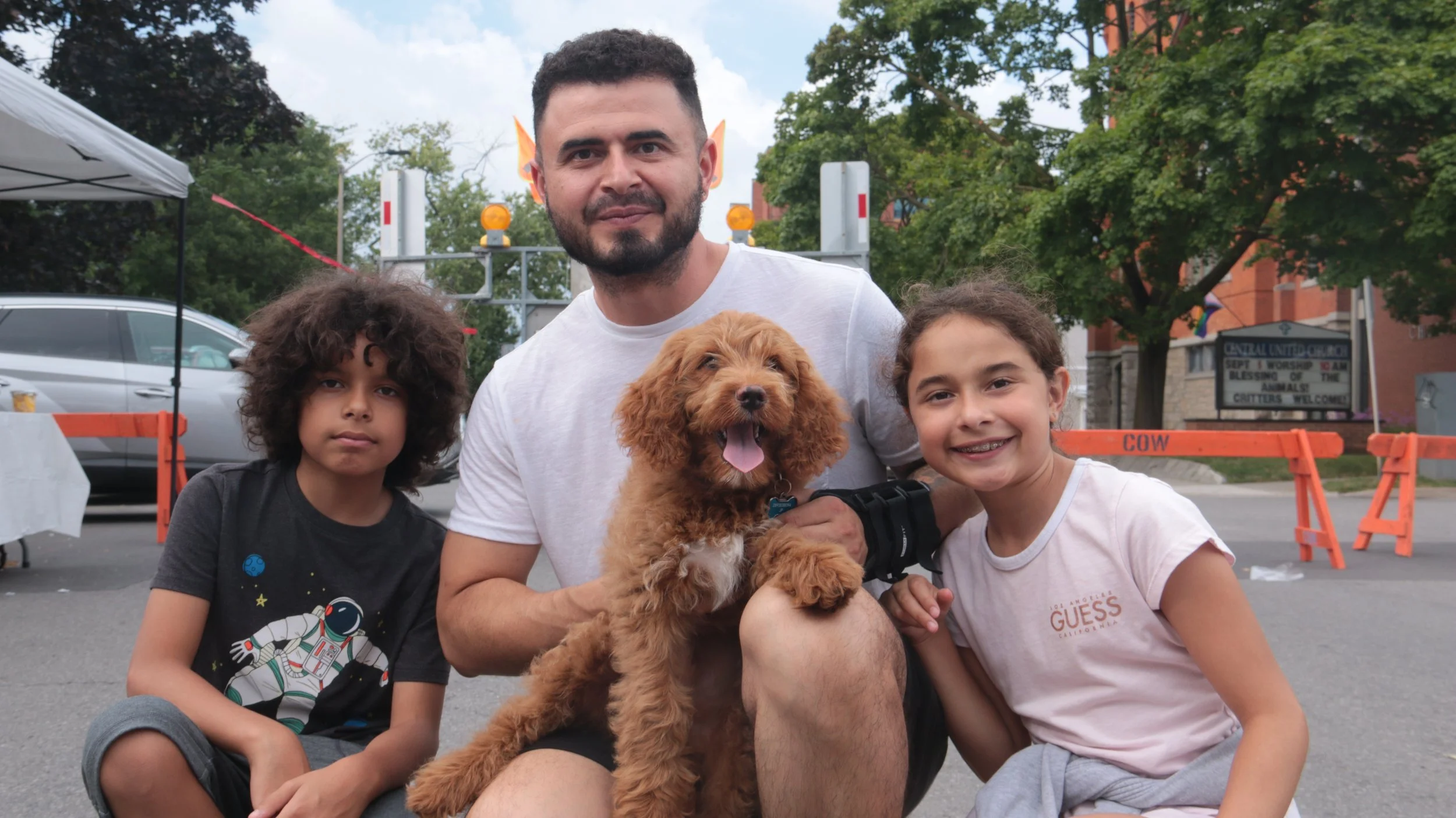 A man with two children and a puppy outdoors near a street with trees, cars, a tent, and orange barricades.