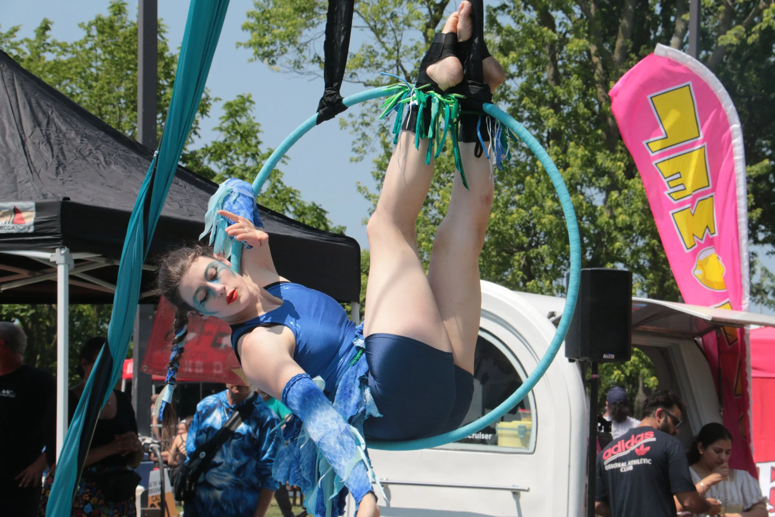 A female aerial performer with makeup and costume hanging upside down in a blue-themed costumes on a hoop at an outdoor event, with people and tents in the background.