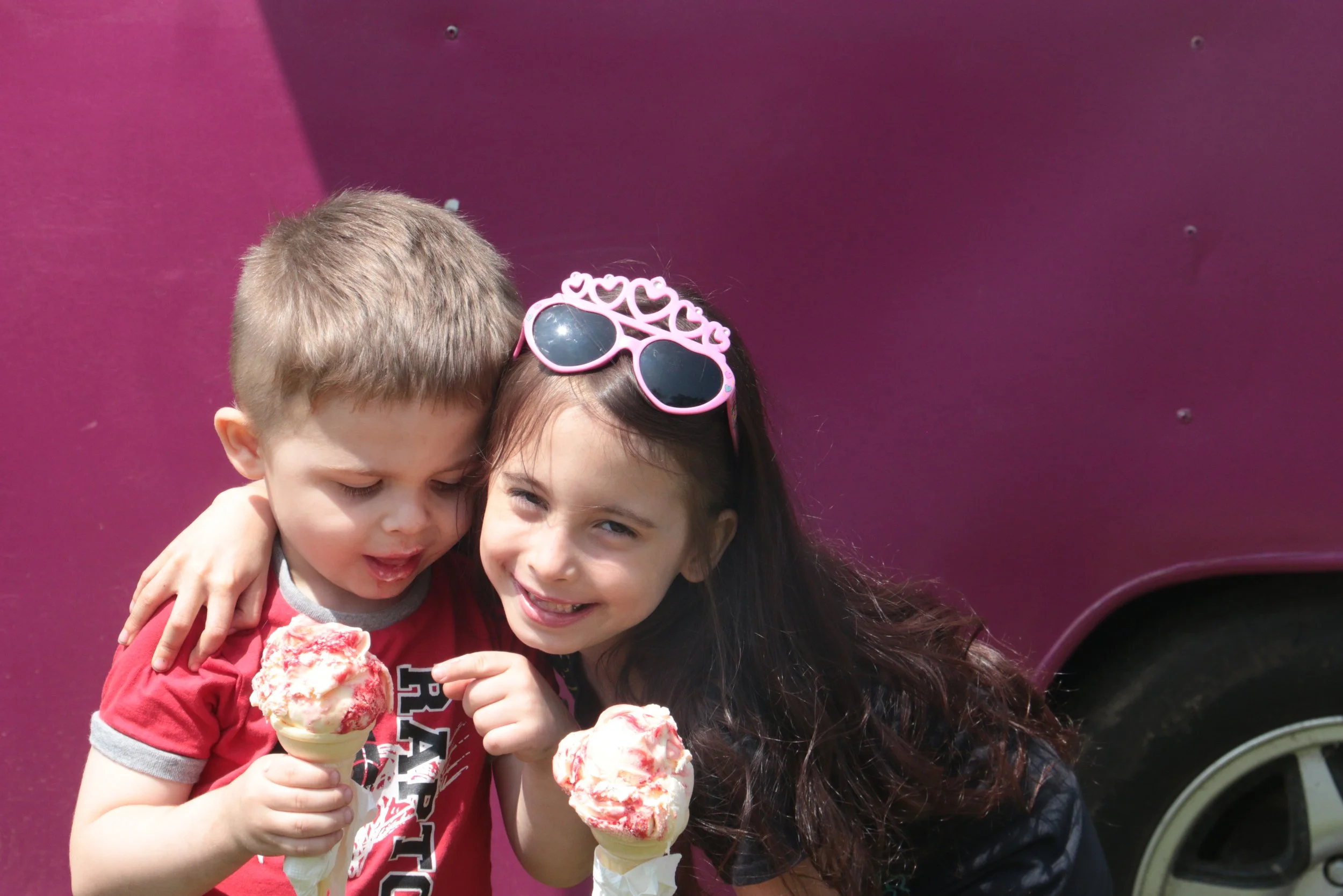 Two children, a boy and a girl, sharing ice cream cones outdoors on a sunny day, with a pink vehicle in the background.