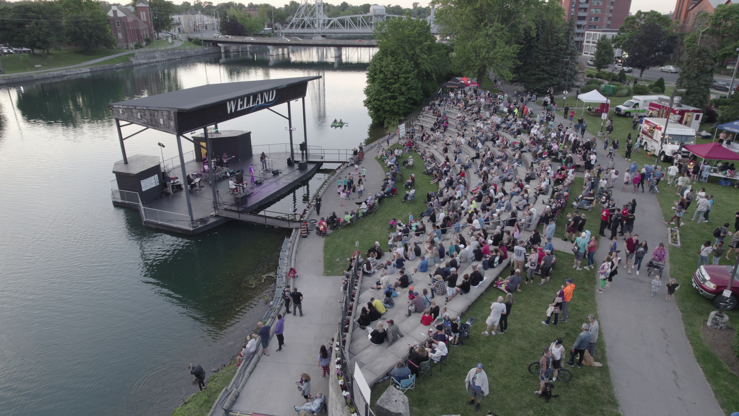 An outdoor concert by a lake with many seated and standing people, a stage with a band performing, food trucks, tents, and trees in the background.
