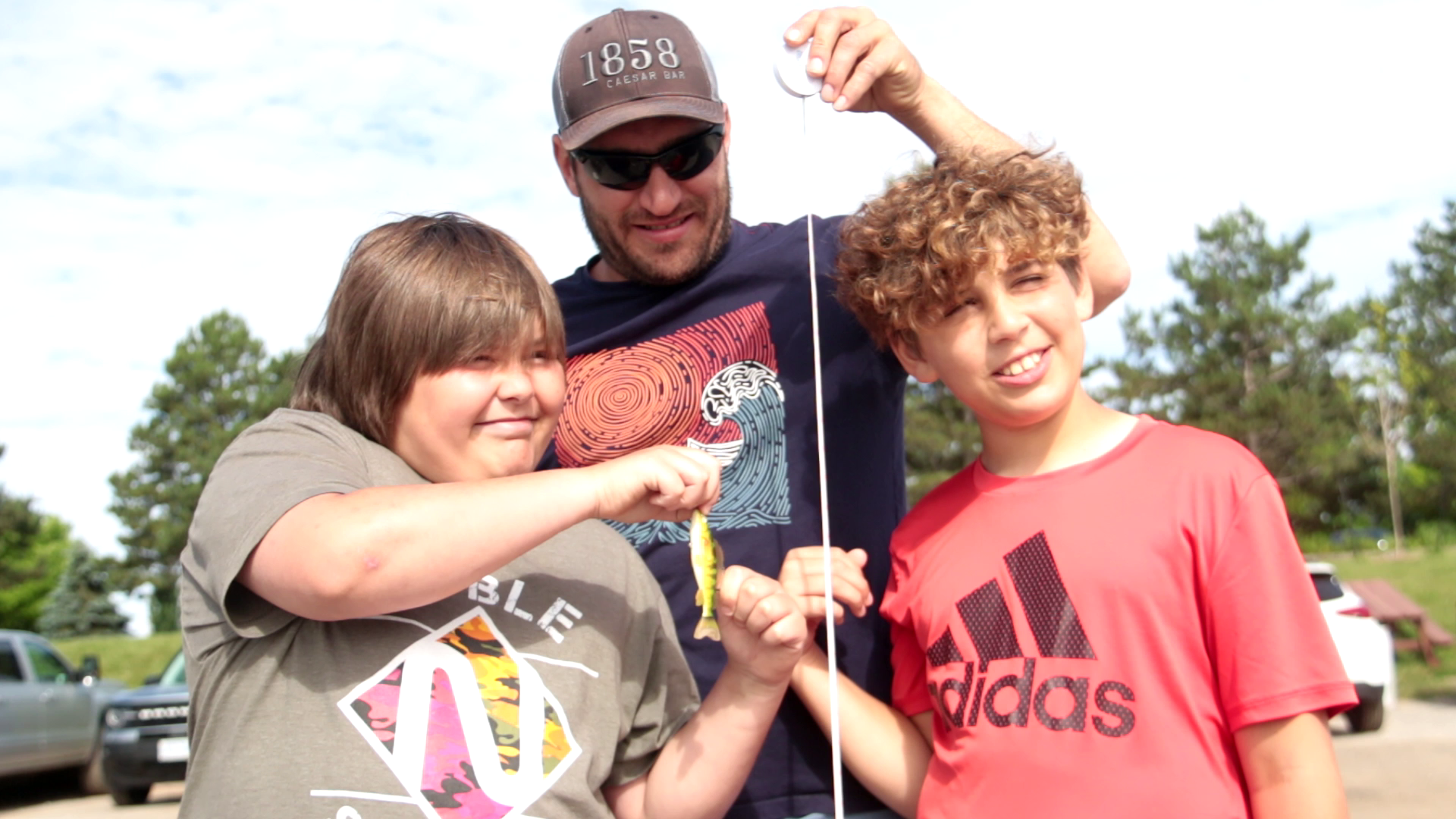 A man with two children holding a small fish on a fishing line outdoors on a sunny day.