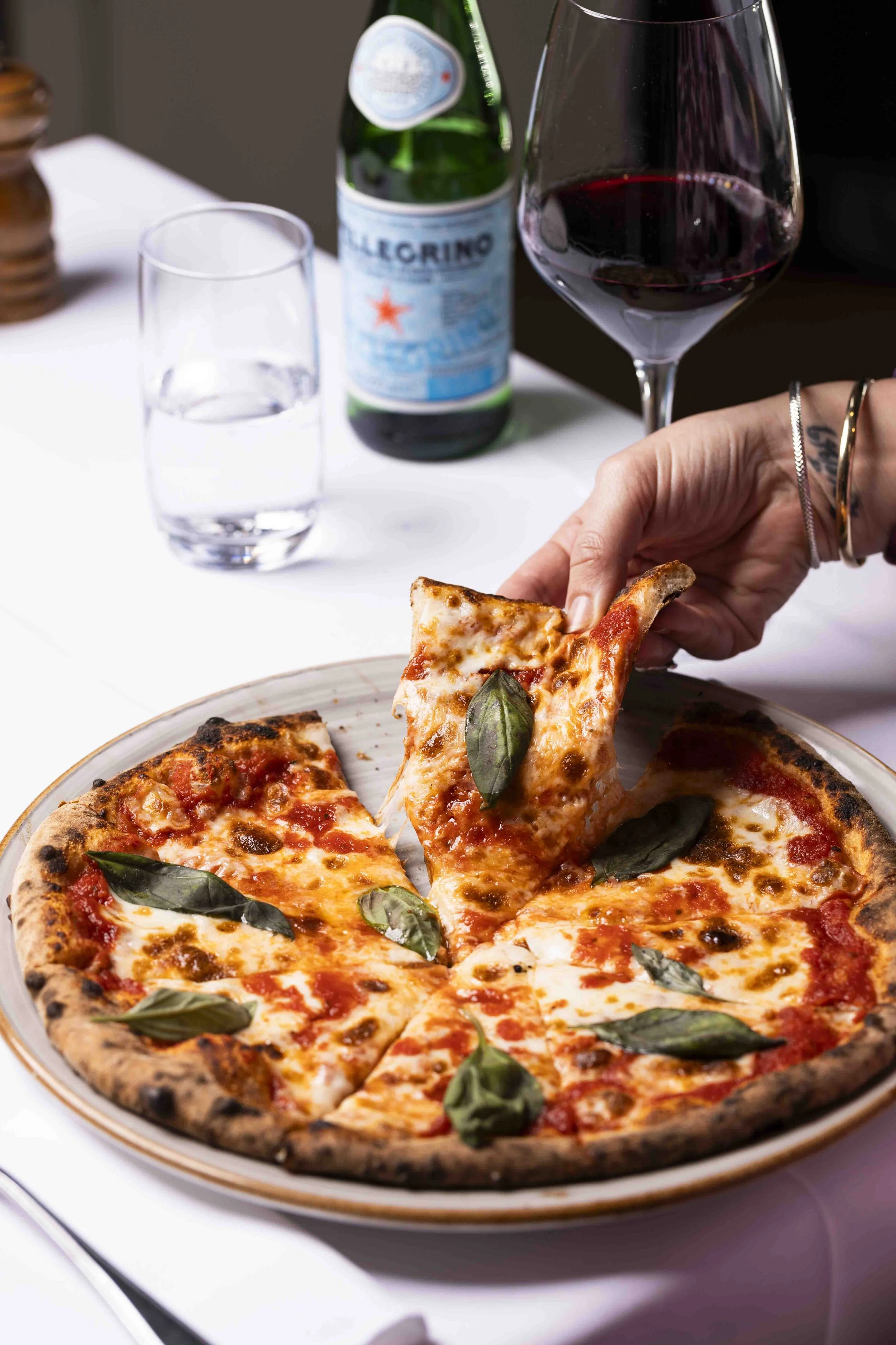 A person is picking up a slice of cheese pizza with basil leaves on it from a pizza tray. There is a glass of red wine, a water glass, and a bottle of San Pellegrino sparkling water on the table.