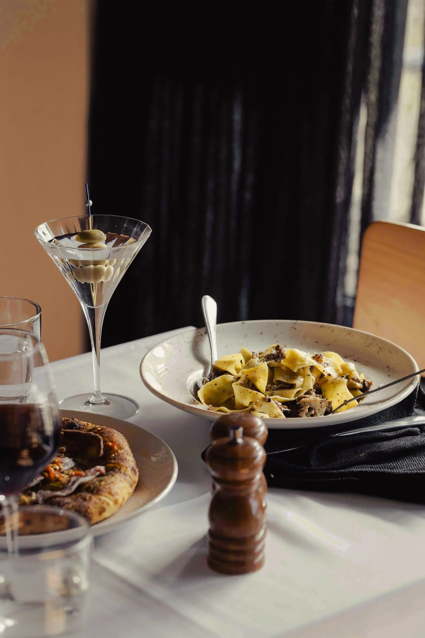 Table setting with a martini glass containing olives, a plate of pasta, a dish with a flatbread, a glass of water, a glass of red wine, and a pepper mill.