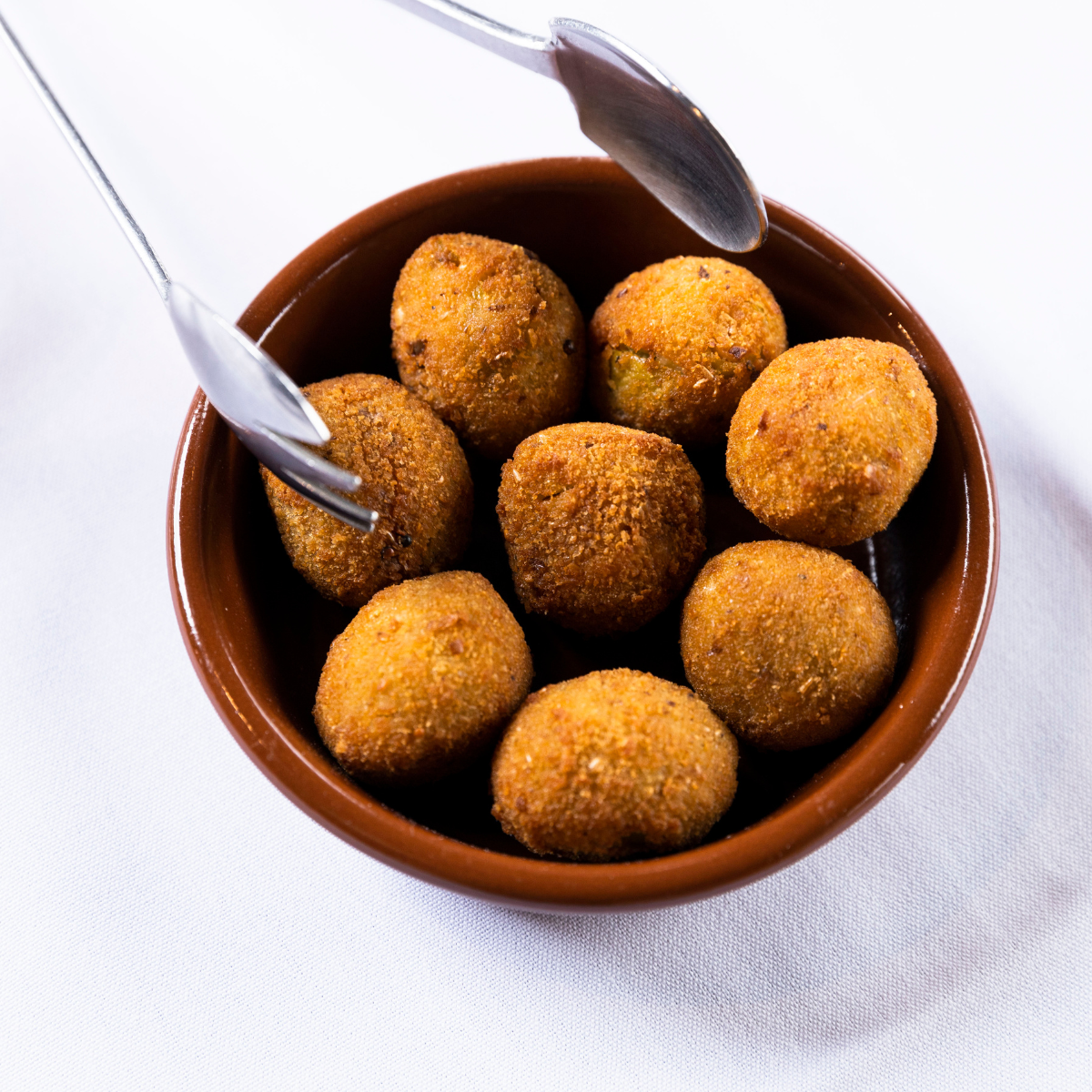 A brown bowl filled with fried arancini balls on white tablecloth, with two metal utensils resting on the bowl.