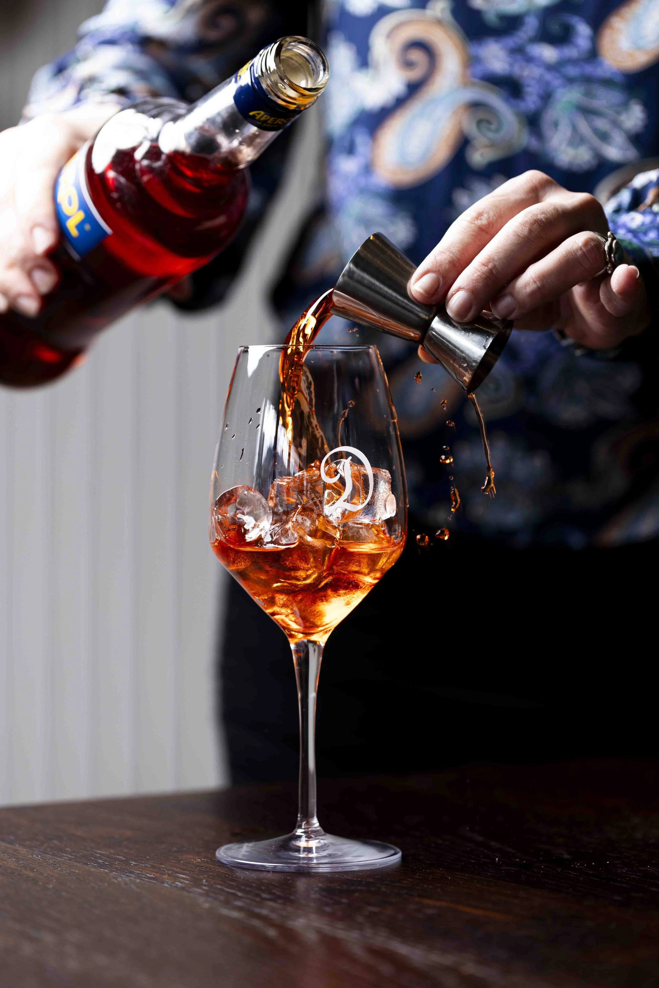 Person pouring Aperol into a glass with ice using a jigger on a wooden table.