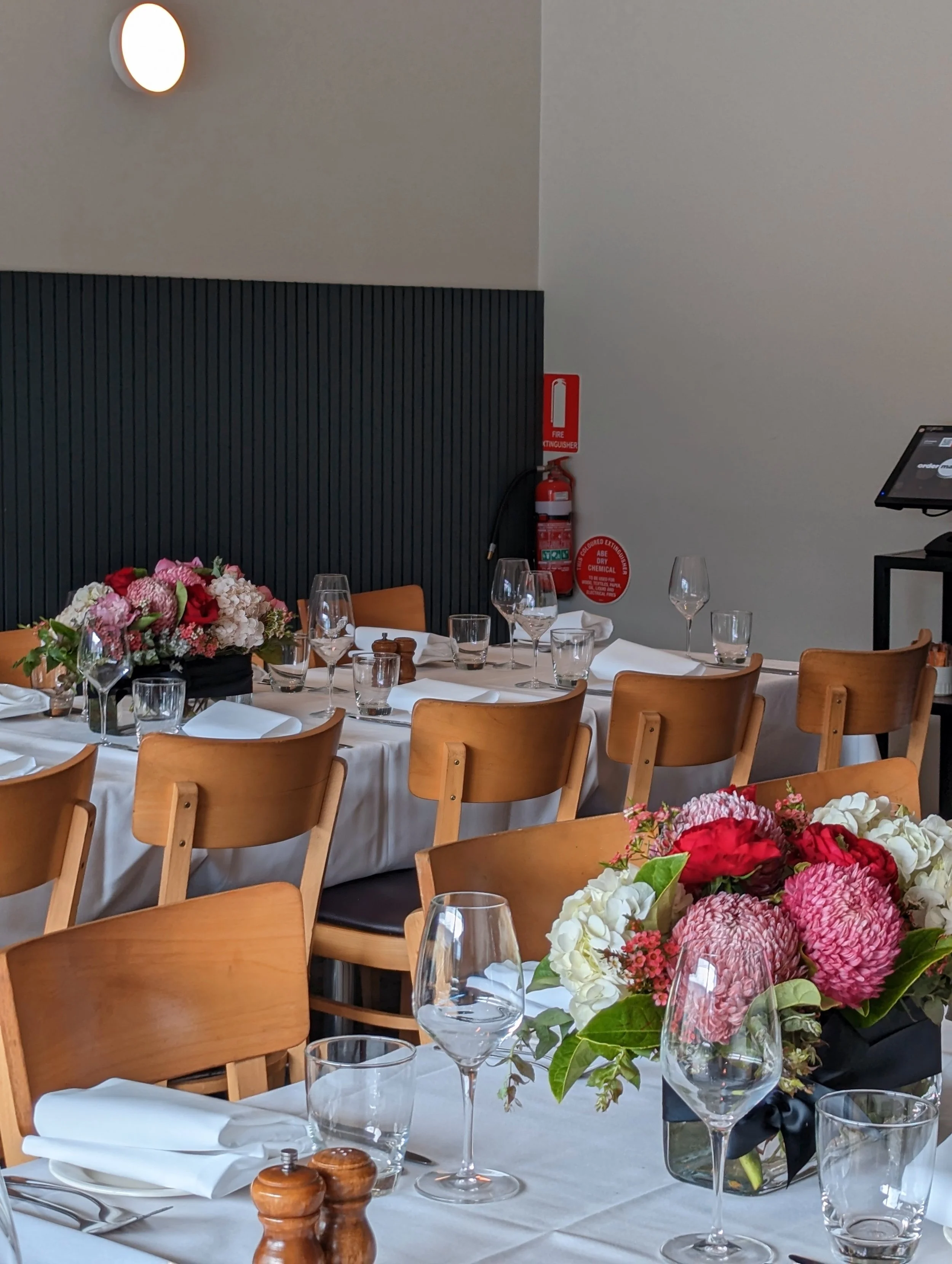 Restaurant interior with wooden chairs and tables set with white tablecloths, glassware, utensils, and flower centerpieces. Fire extinguisher visible on the wall. Soft lighting from a wall lamp.
