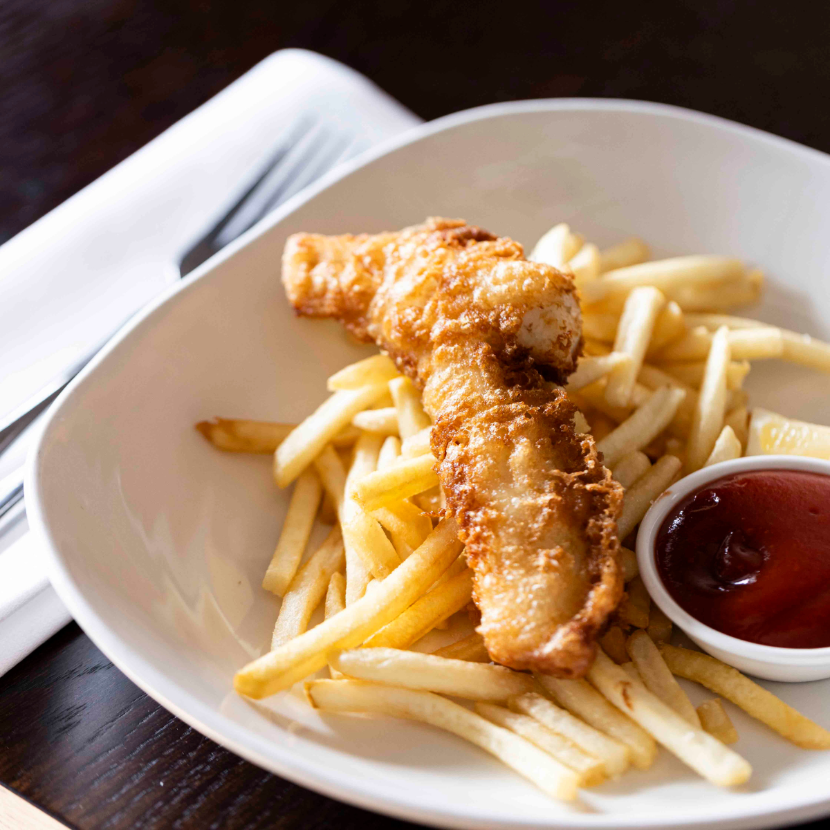 Fried fish fillet on a bed of French fries with a side of ketchup in a small dish.