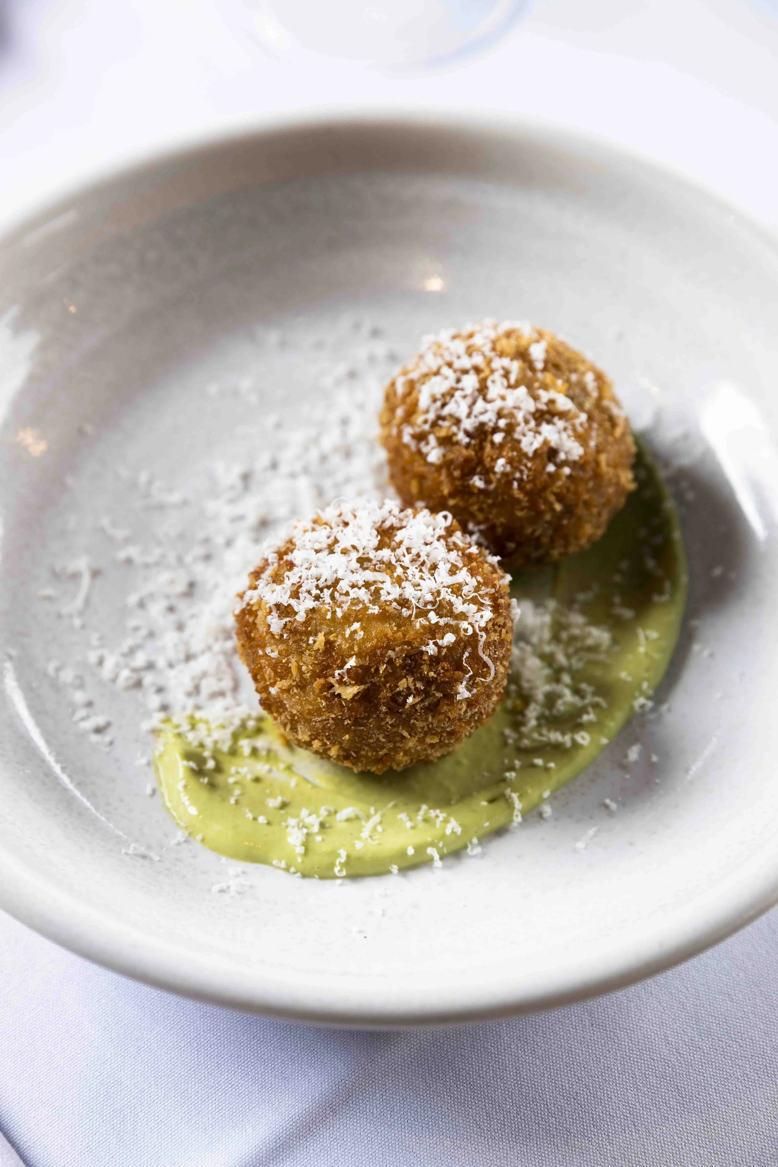 Two fried arancini balls garnished with grated cheese, placed on a green sauce in a white bowl.