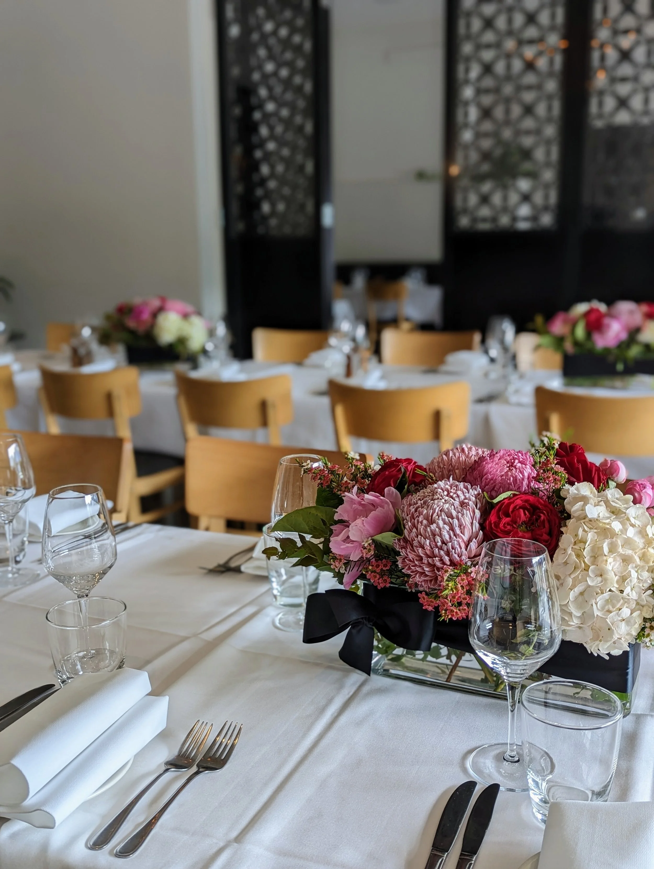 Elegant dining table set with white tablecloths, floral centerpieces, wine glasses, and cutlery in a formal restaurant setting.