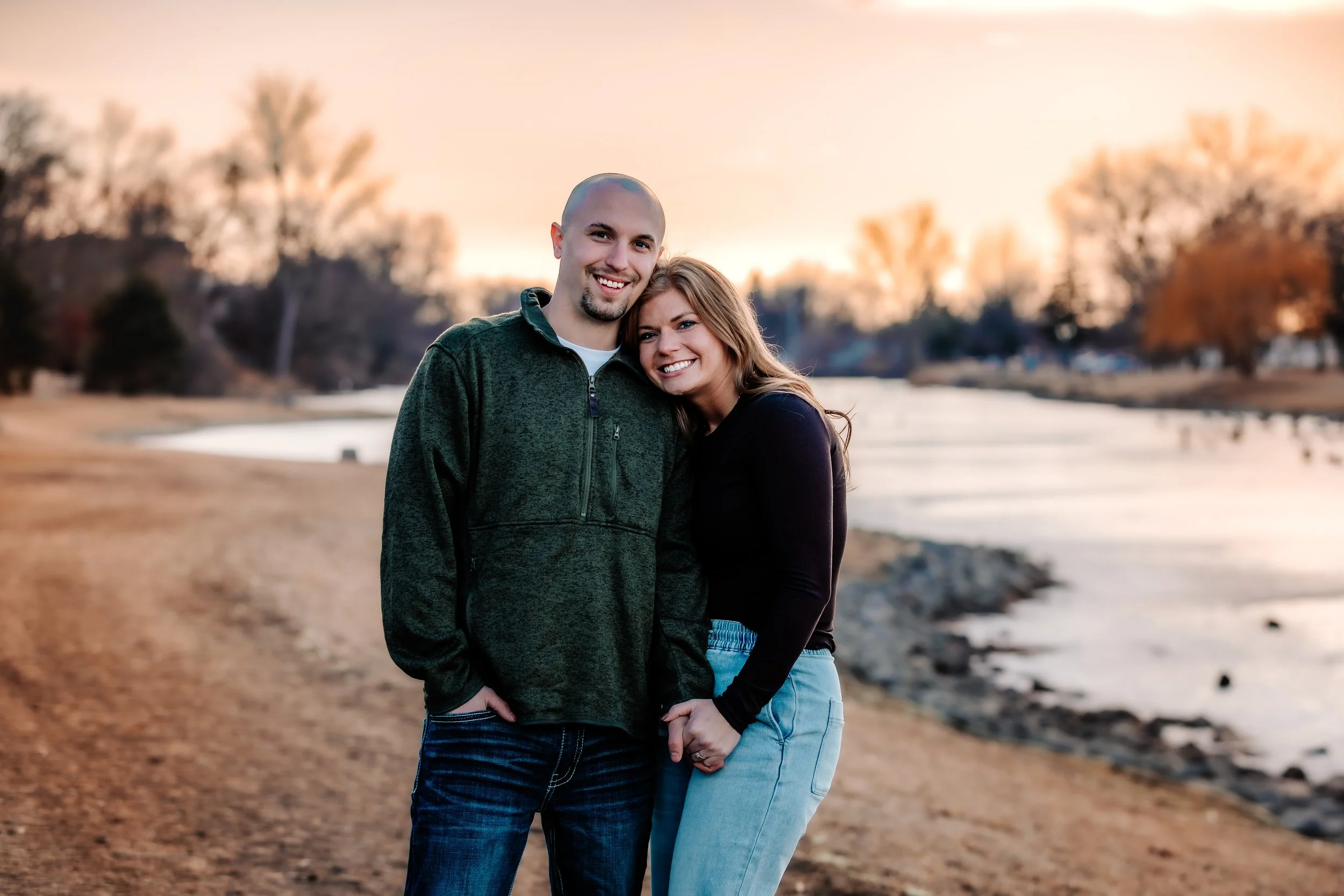 A smiling couple standing near a river at sunset, holding hands, with trees in the background at Terrace Park in Sioux Falls, South Dakota.