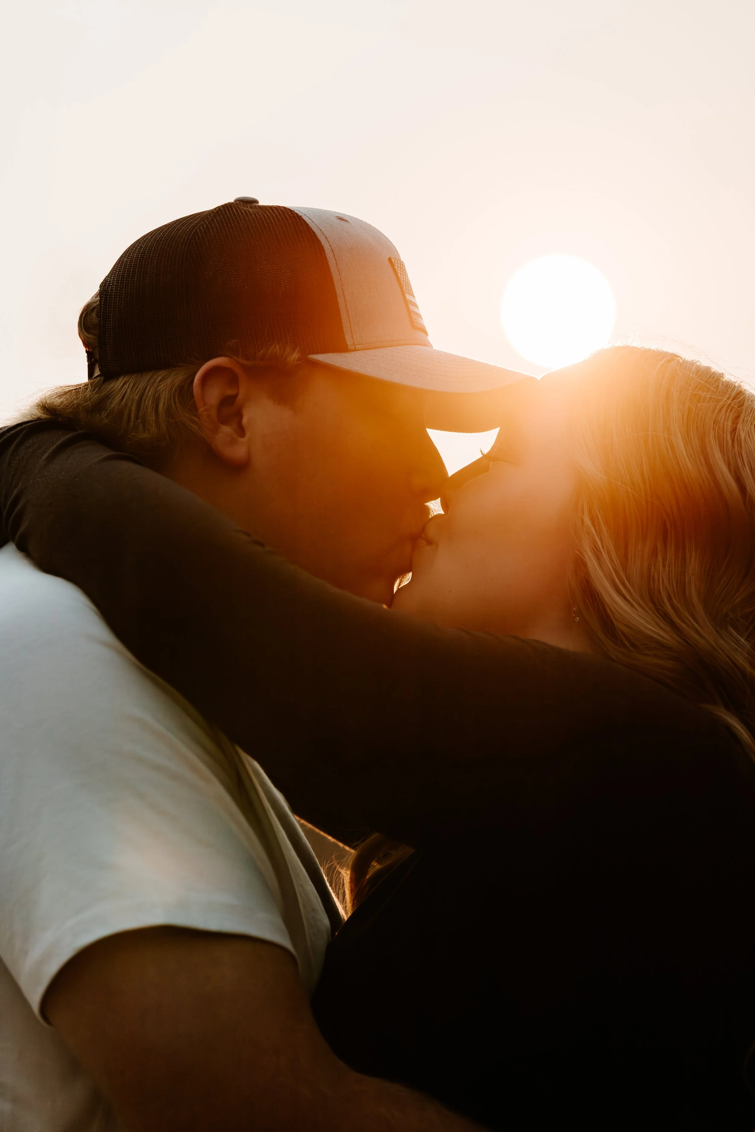 A couple kisses during sunset, with the man wearing a cap and the sun shining in the background in Downtown Sioux Falls, South Dakota.