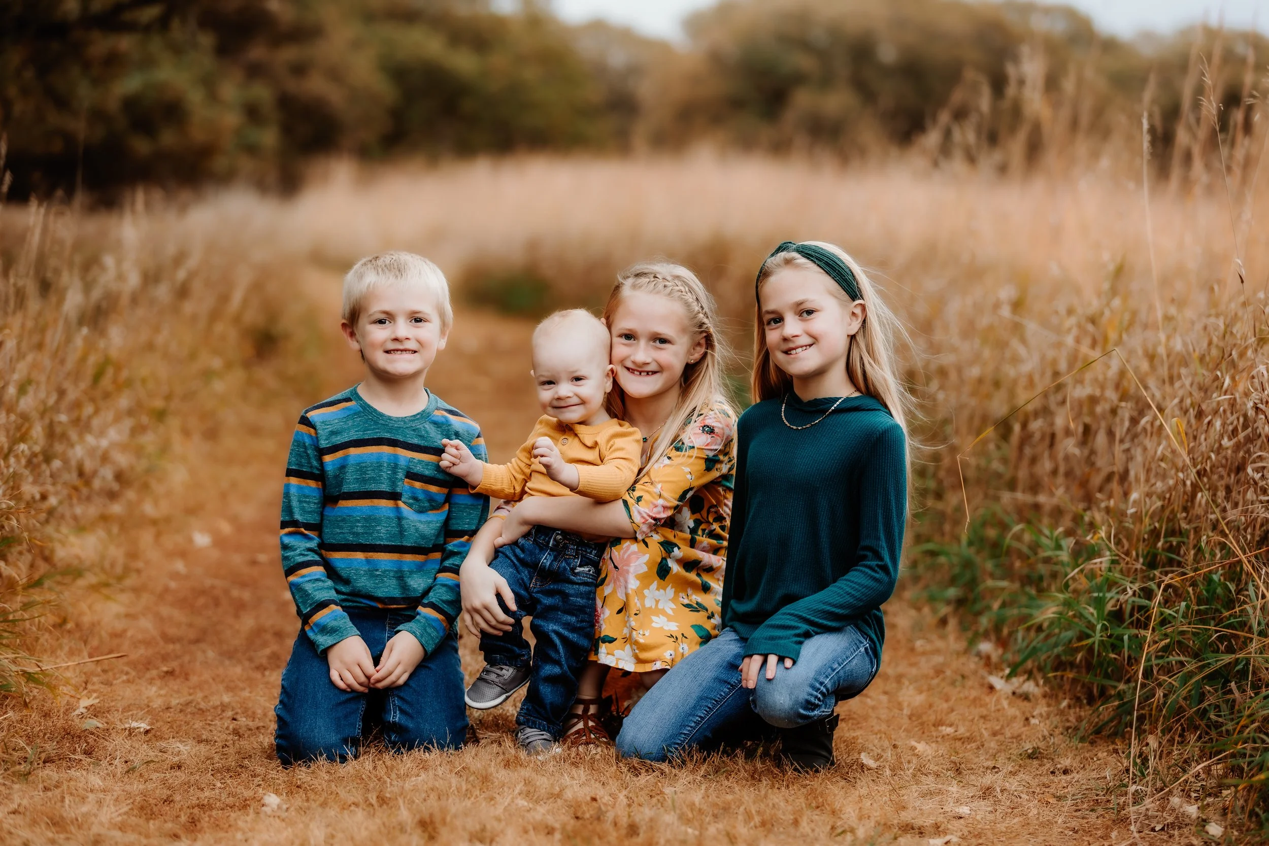 Four children, two boys and two girls, sitting on a dirt path through a field of tall grass and trees in the background. They are smiling and dressed in fall clothing in Sioux Falls, South Dakota at Good Earth State Park.