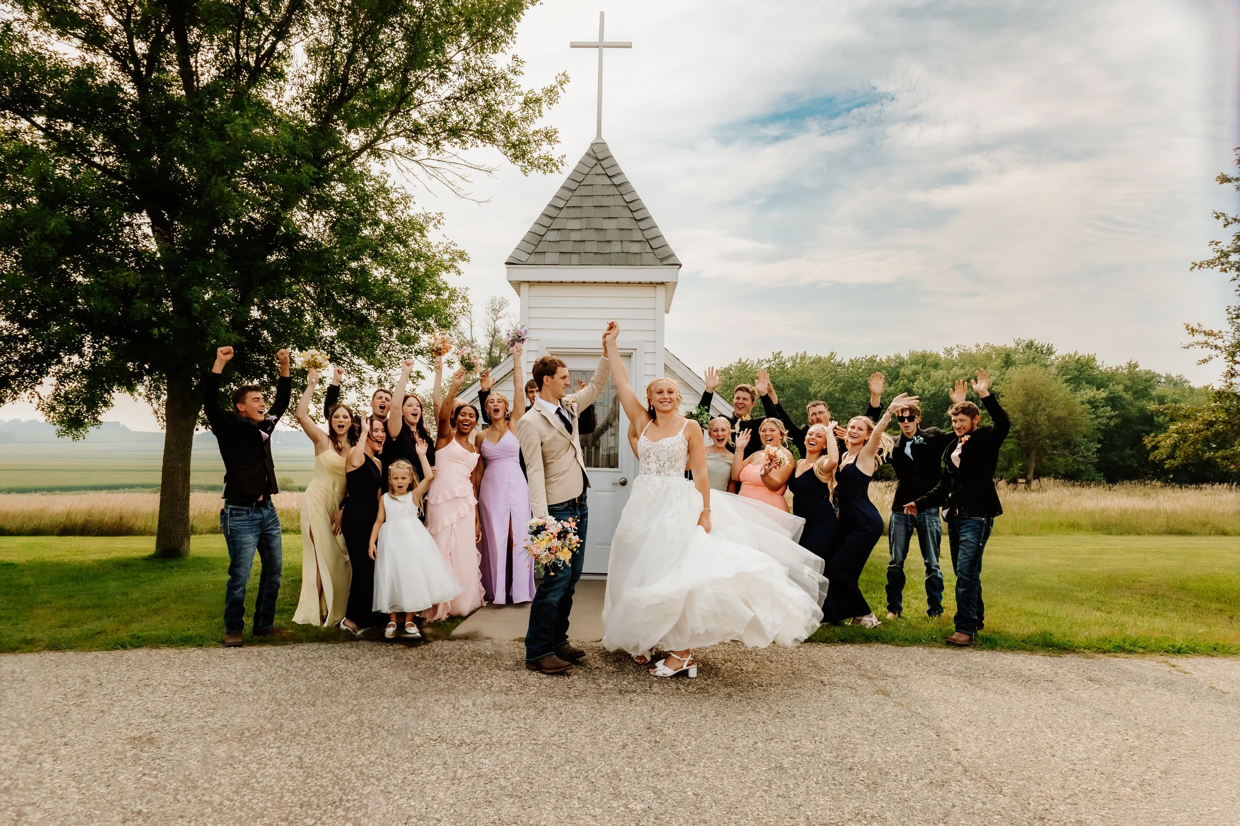 A bride and groom celebrating with friends and family outside a small white chapel, with trees and a field in the background, on a partly cloudy day in Luverne, MN.