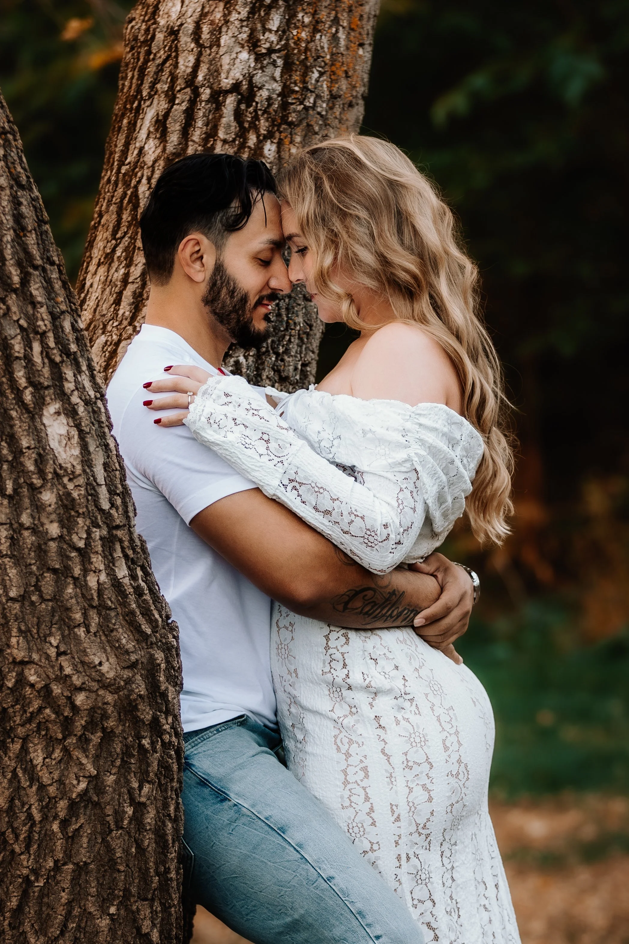 A couple embraces affectionately outdoors, leaning against a tree, with their foreheads touching and eyes closed at Mary Jo Wegner Arboretum in Sioux Falls, South Dakota.