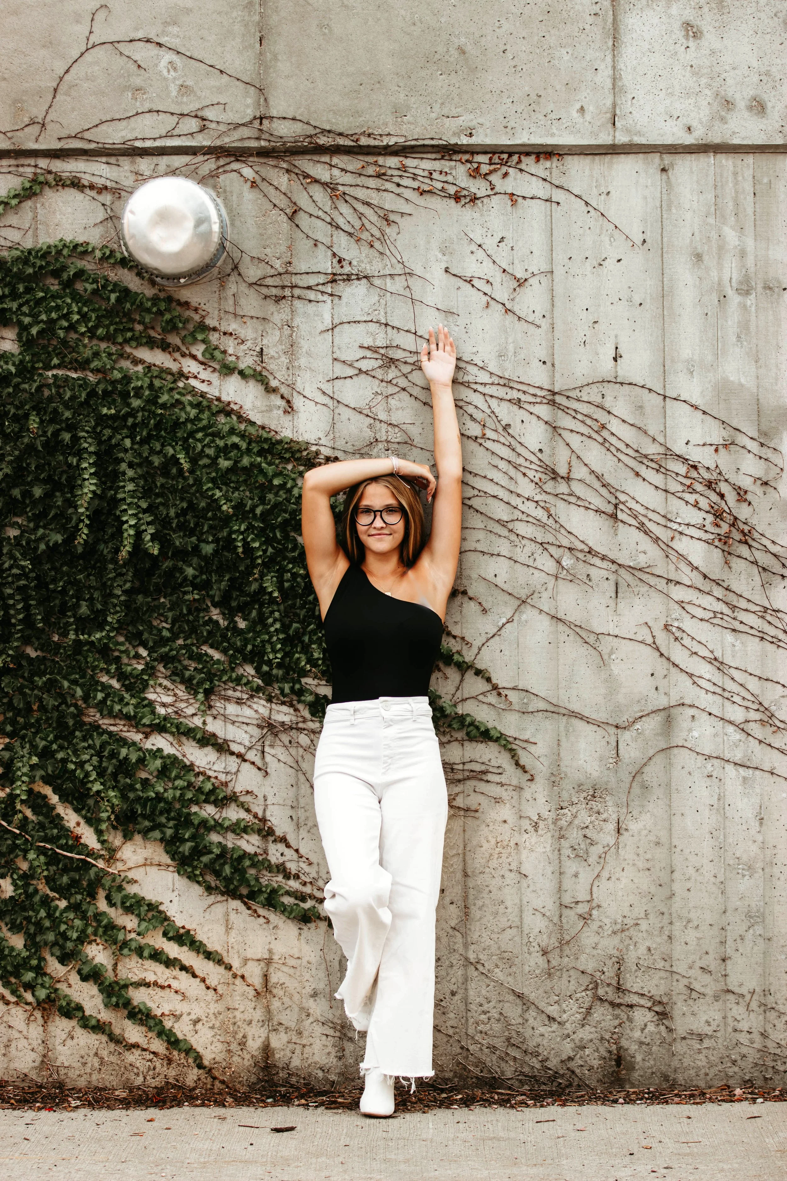 A young woman with glasses wearing a black one-shoulder top and white jeans, leaning against a concrete wall with climbing vines, raising one arm in the air in Downtown Sioux Falls, South Dakota.