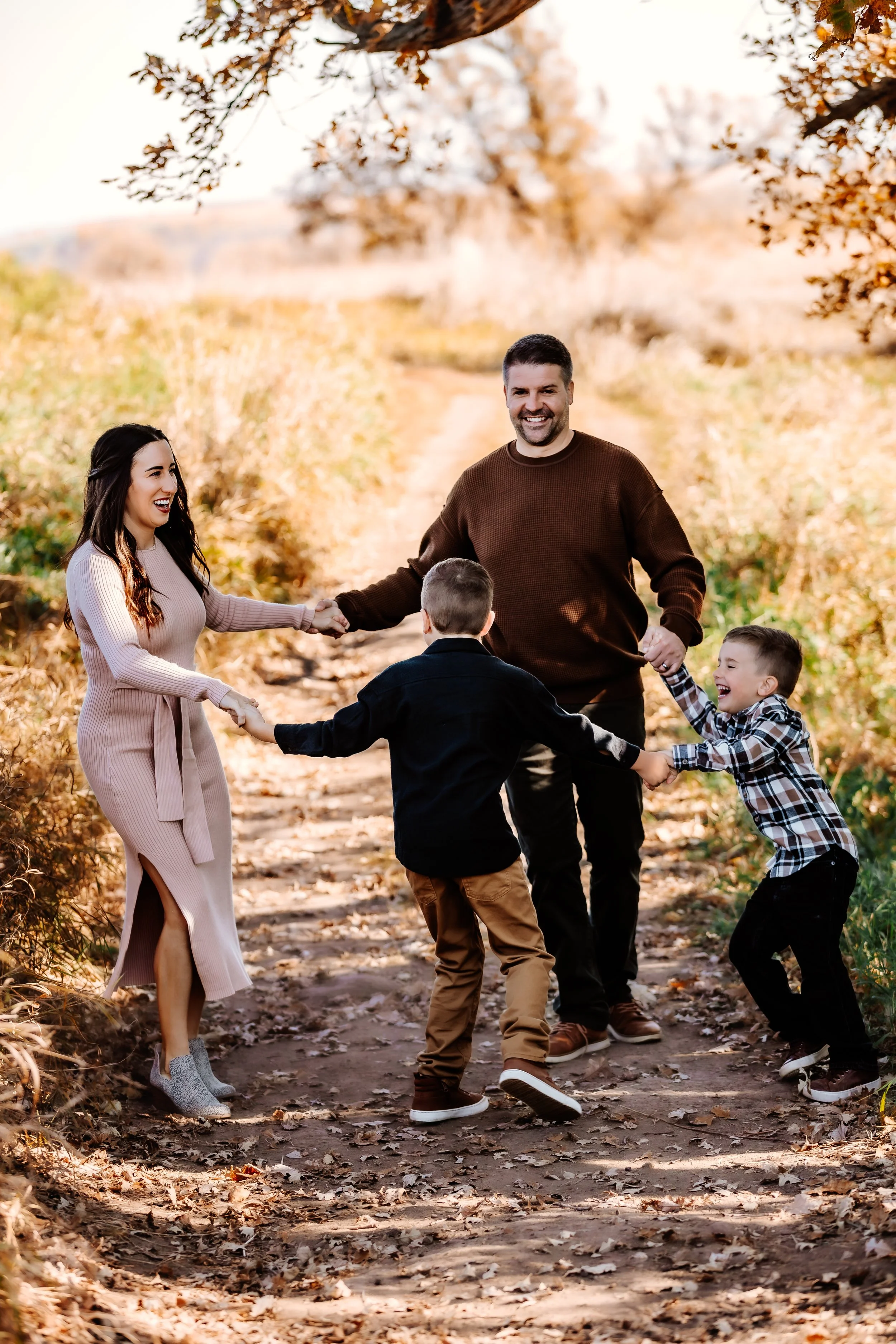 A smiling family of four holding hands in a circle while standing on a fall leaf-covered path in a park, with trees and autumn foliage in the background in Sioux Falls, South Dakota at Good Earth State Park.