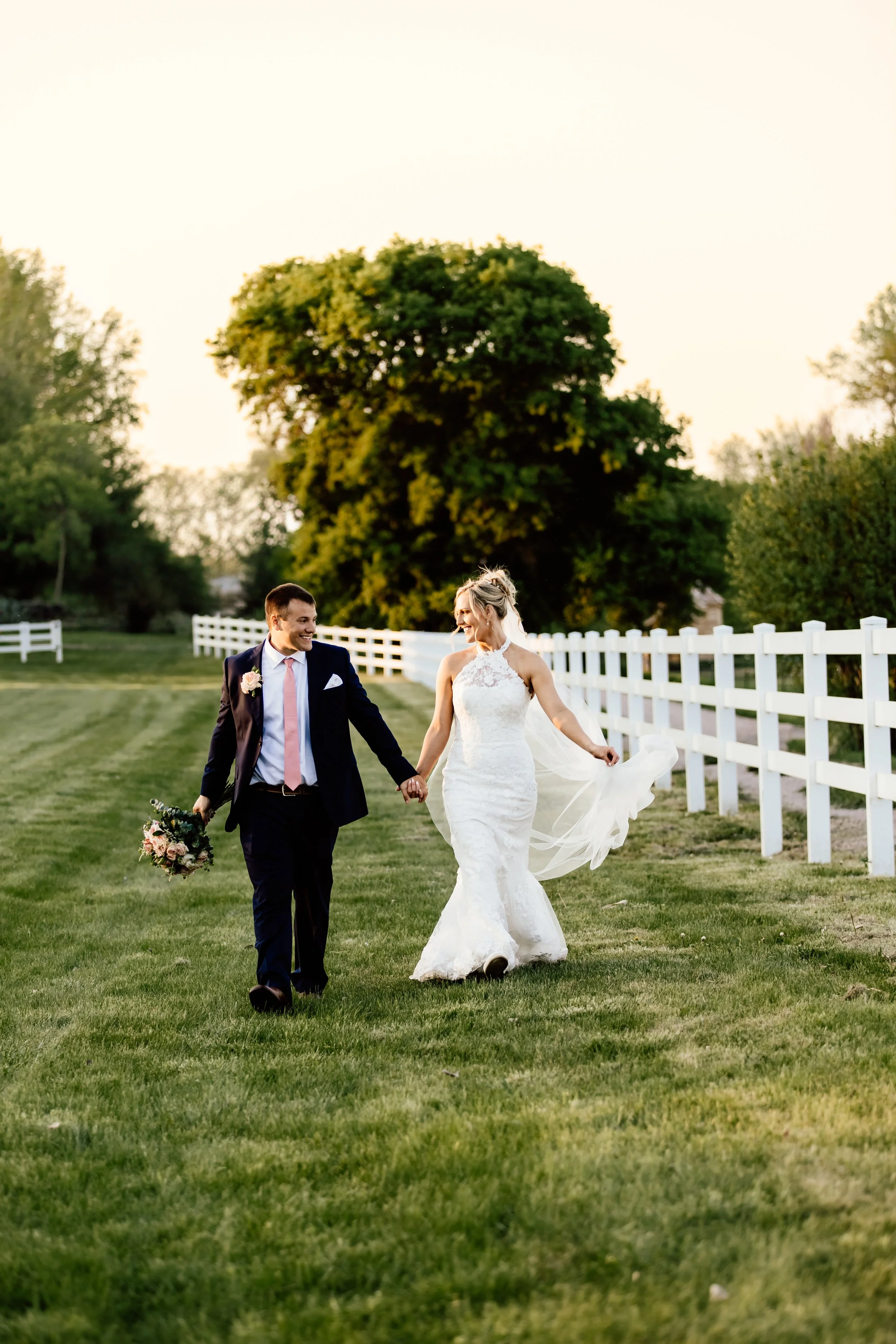 A newlywed couple walking hand-in-hand on a grassy field, with the bride in a white lace wedding dress and the groom in a dark suit holding a bouquet, surrounded by a white fence and green trees at sunset at Laurel Ridge in Sioux Falls SD.
