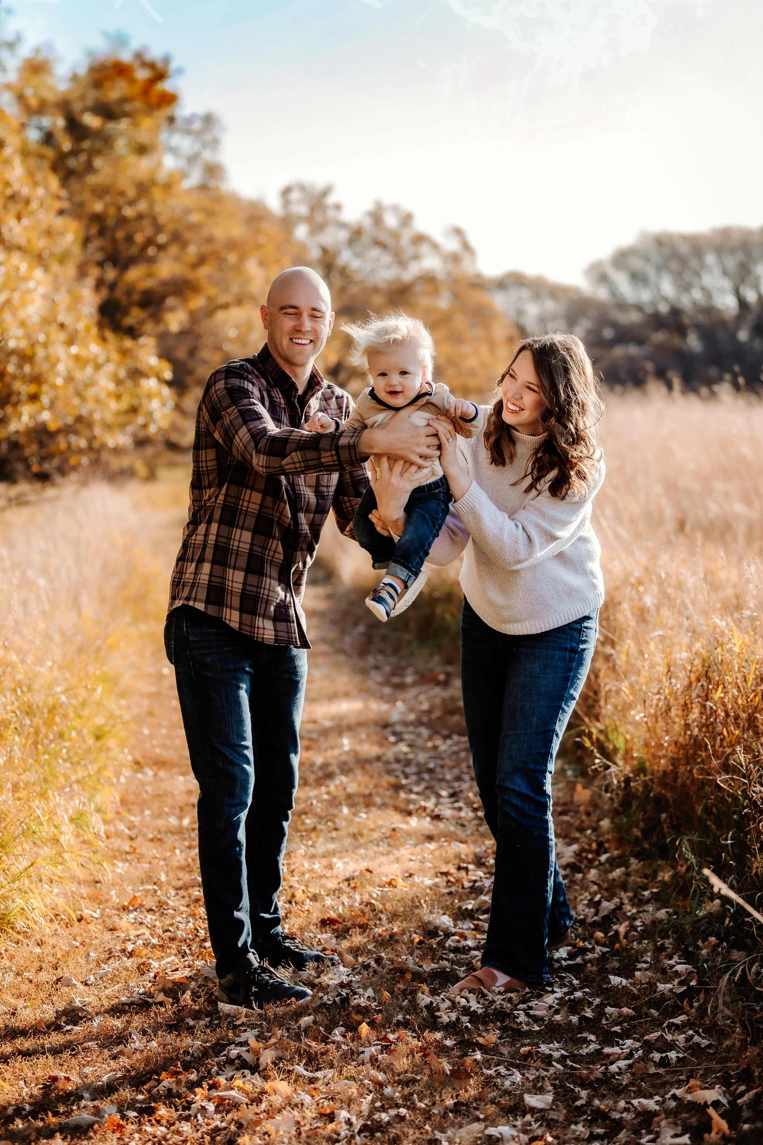 A family of three having fun outdoors in an autumn setting with fall foliage, with a man and woman smiling and holding a young child between them in Sioux Falls, South Dakota at Good Earth State Park.