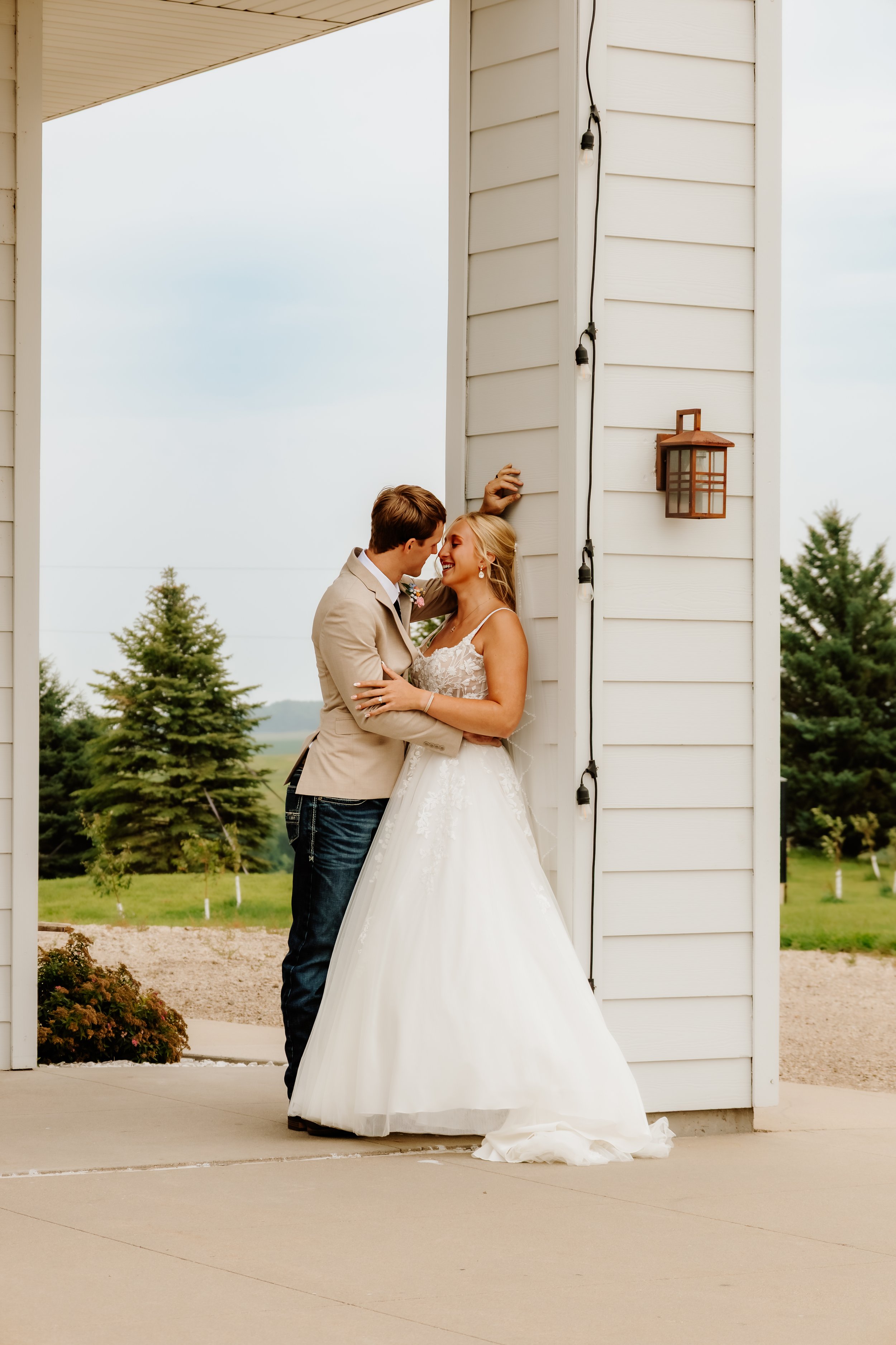 A bride and groom share a moment outside near a barn, with the groom leaning against the wall and the bride embracing him in Luverne, MN.