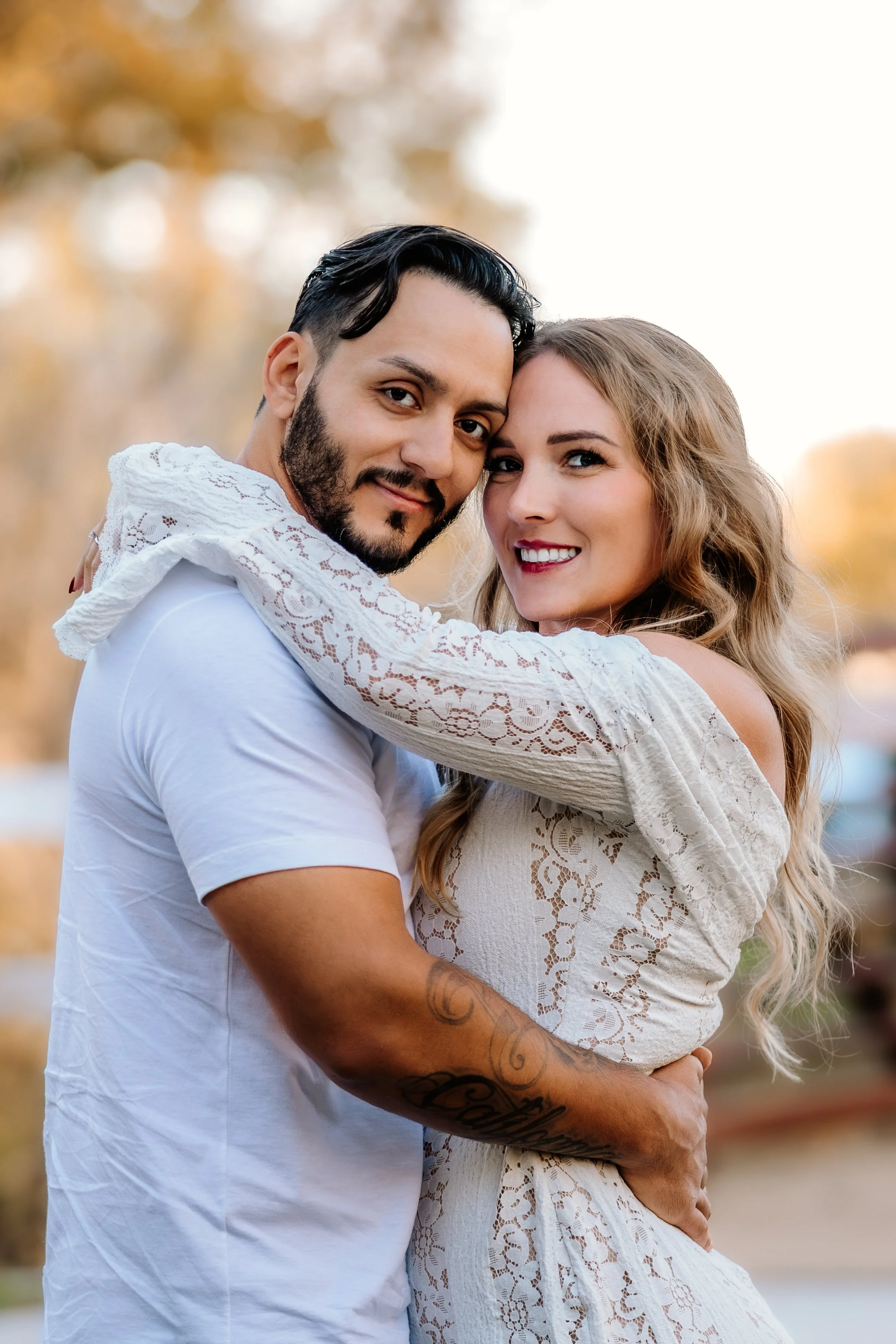 A happy couple embracing outdoors during sunset, with the man wearing a white t-shirt and the woman in a lace dress at Mary Jo Wegner Arboretum in Sioux Falls, South Dakota.