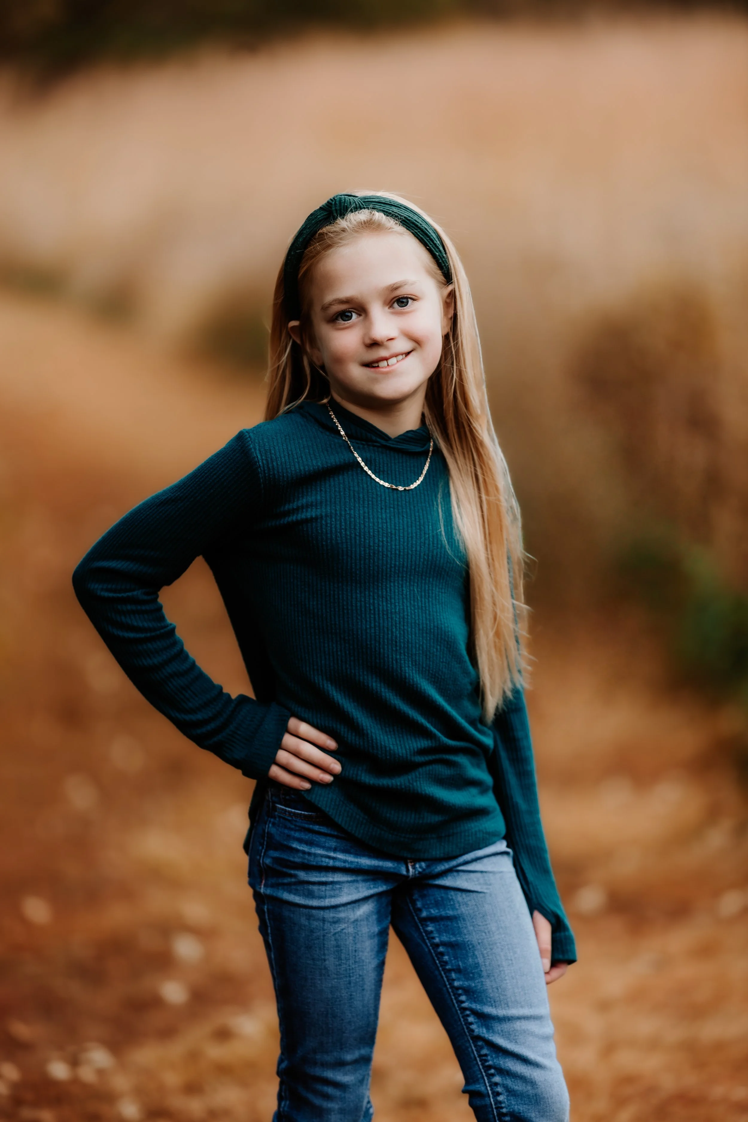 Young girl with long blonde hair, wearing a dark green sweater, jeans, and a headband, standing outdoors with one hand on her hip, smiling at the camera in Sioux Falls, South Dakota at Good Earth State Park.