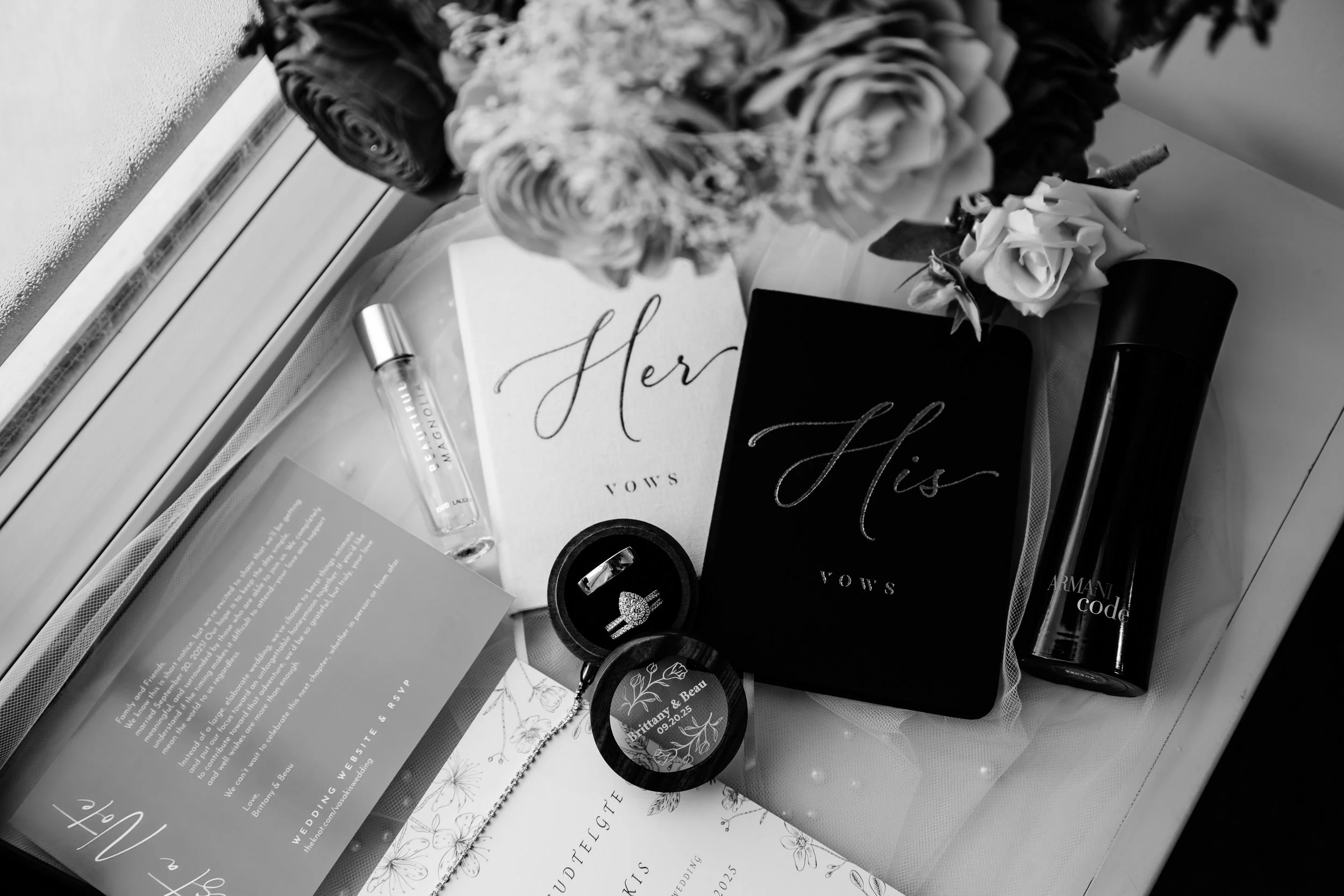 Wedding vow booklets labeled "Her" and "His", a small bouquet of roses, wedding rings, a wedding invitation, a perfume bottle, a small jewelry box with an engagement ring, and a wedding hashtag sign on a bed in Sioux Falls, South Dakota.