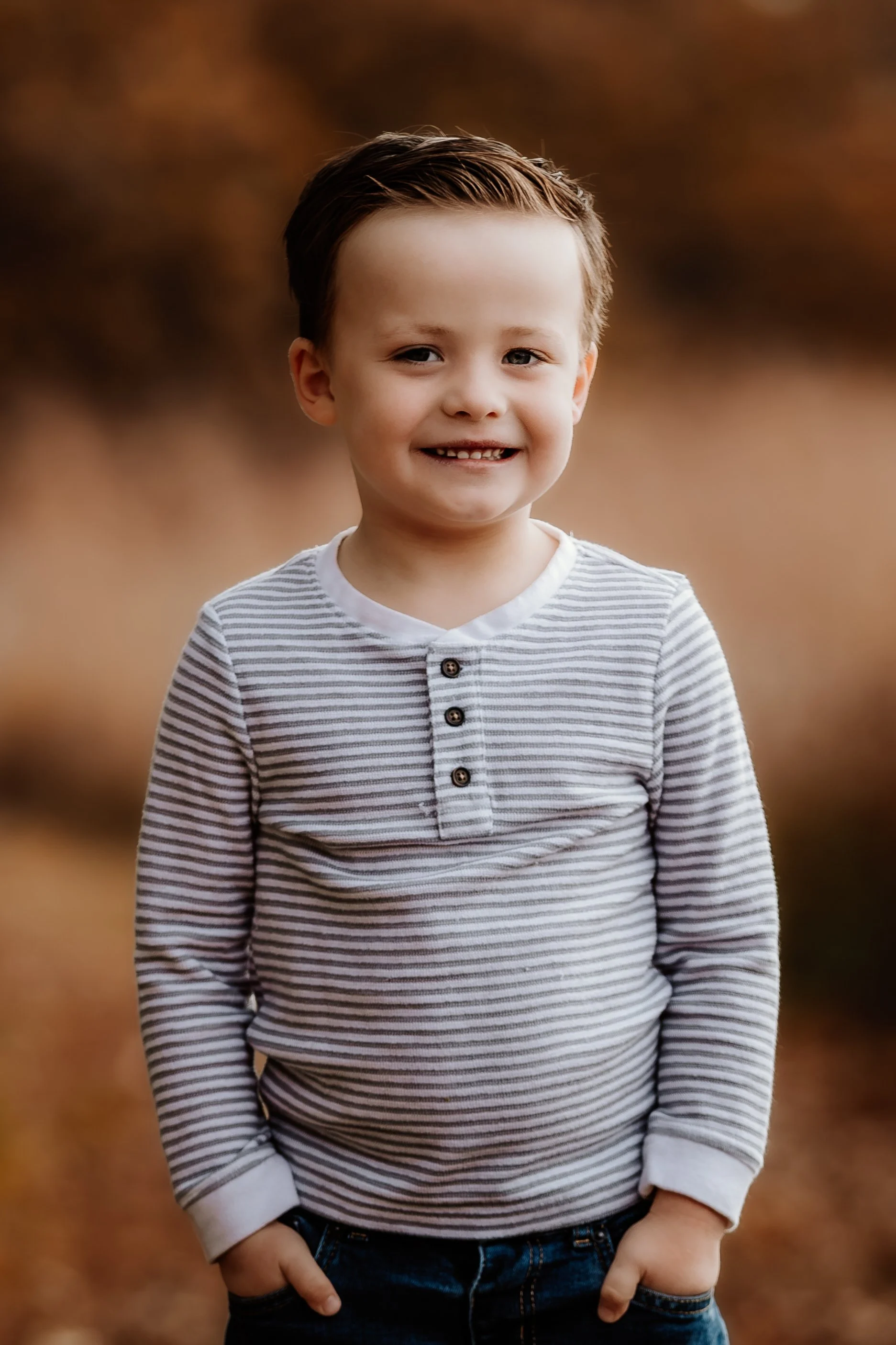 A young boy with short brown hair smiling outdoors, wearing a striped long-sleeve shirt with buttons and dark jeans in Sioux Falls, South Dakota at Good Earth State Park.