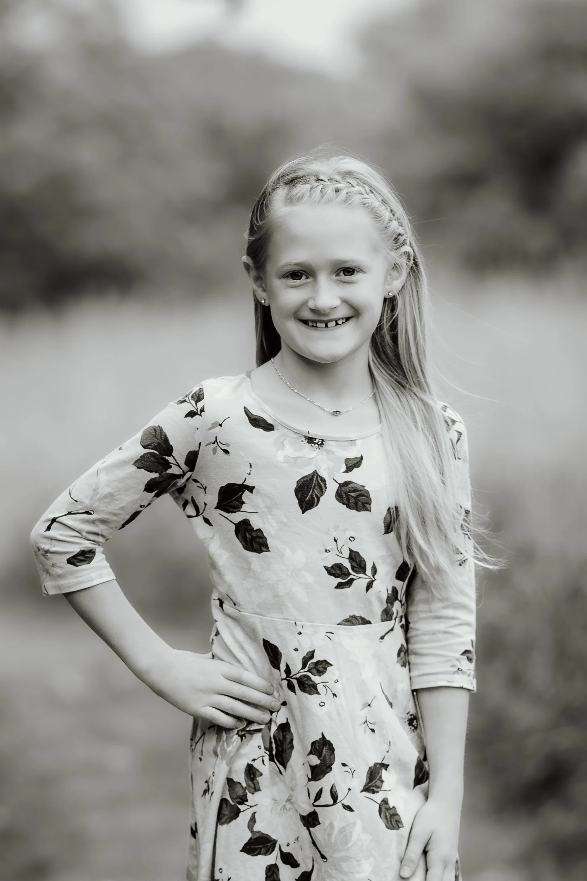 A young girl with long hair, smiling and standing outdoors with her hand on her hip, wearing a floral dress in Sioux Falls, South Dakota at Good Earth State Park.
