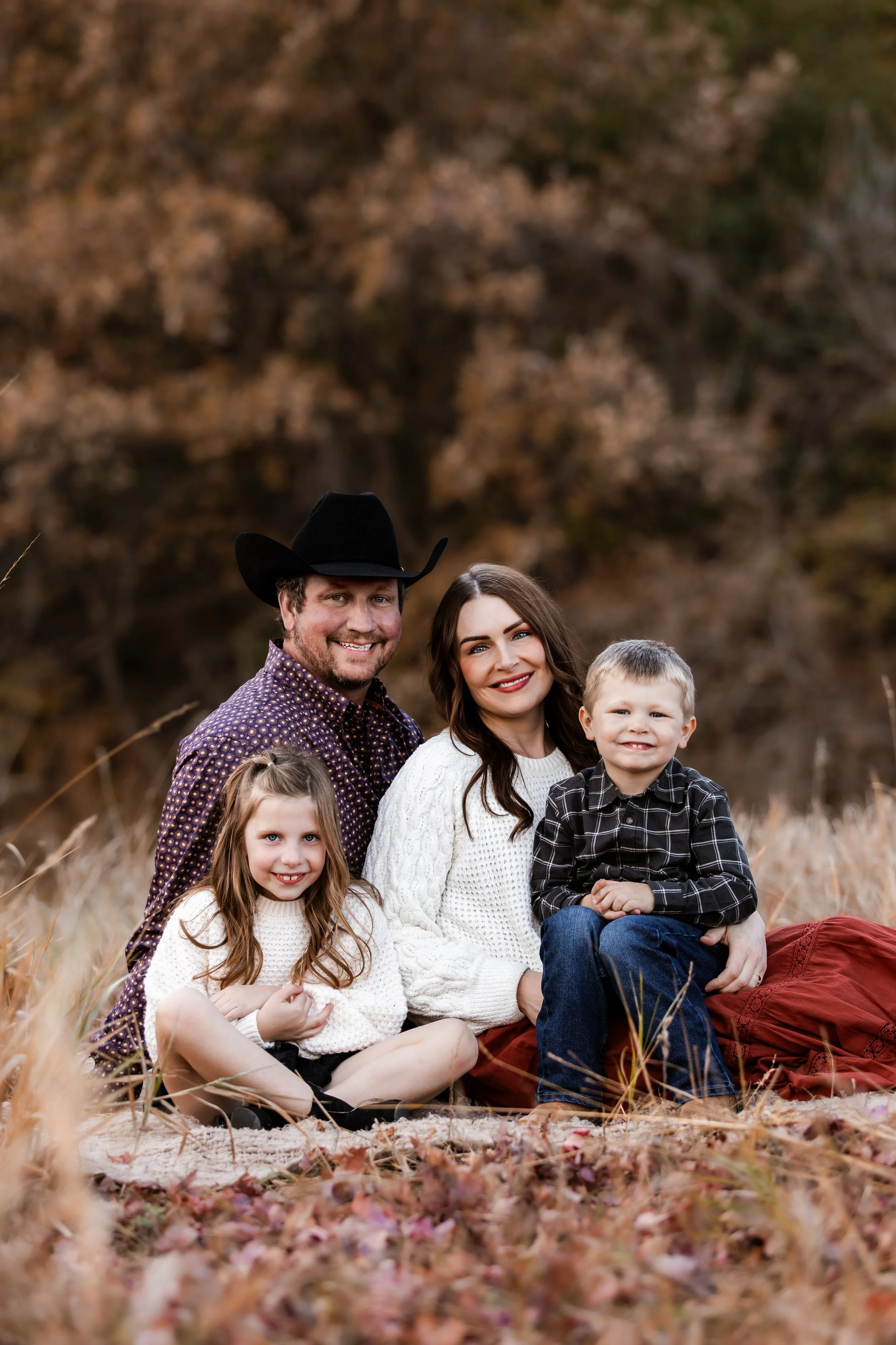 Family of four, including father, mother, daughter, and son, sitting outdoors on autumn foliage with trees in the background at Legacy Park in Sioux Falls, South Dakota.