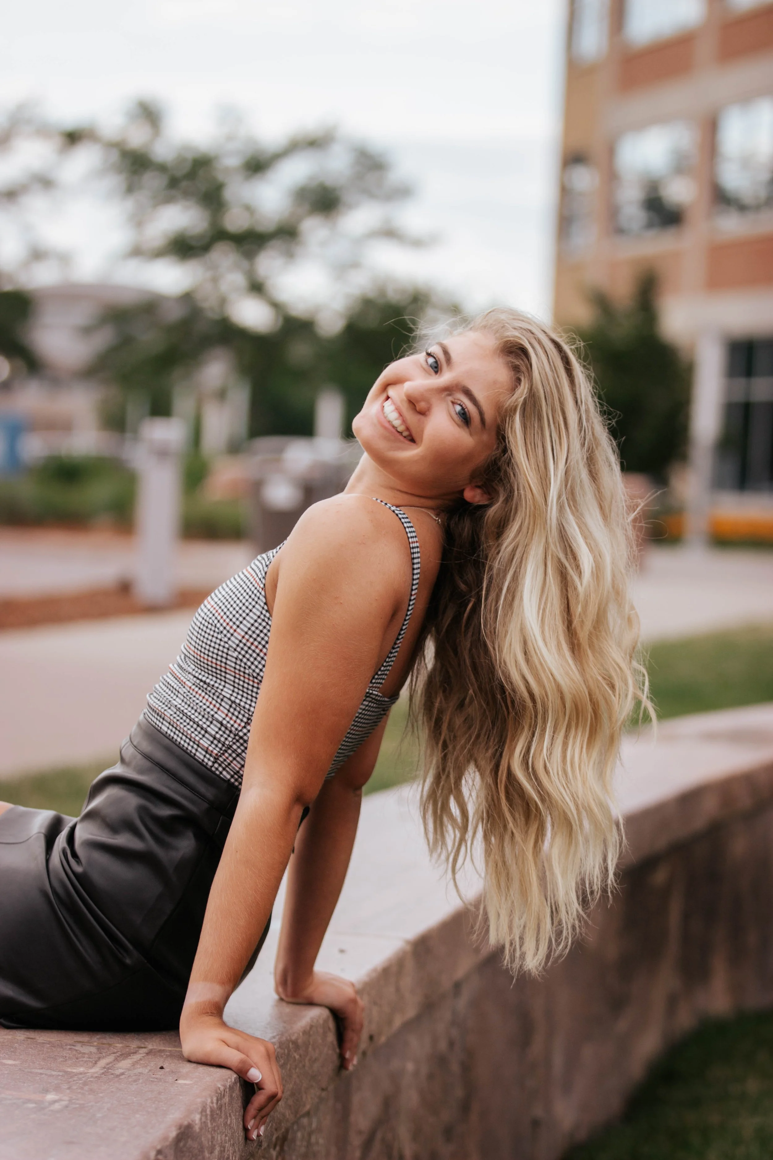 A smiling young woman with long wavy blonde hair is leaning back on a stone wall outdoors, with a blurred background of trees and a building in Downtown Sioux Falls, South Dakota.