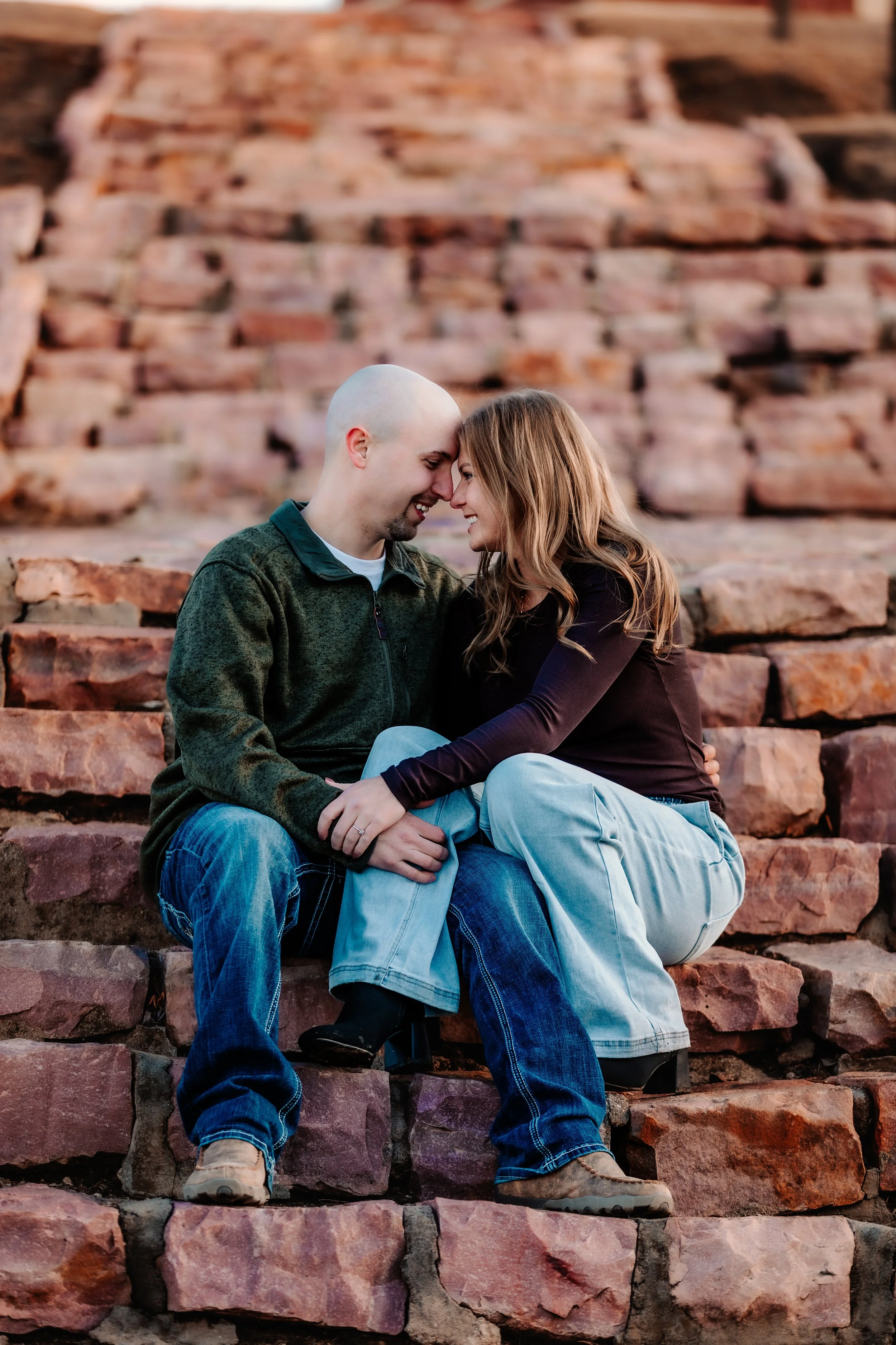 A man and woman sitting close together on stone steps, touching foreheads and smiling at each other. Background of red stone stairs at Terrace Park in Sioux Falls, South Dakota.