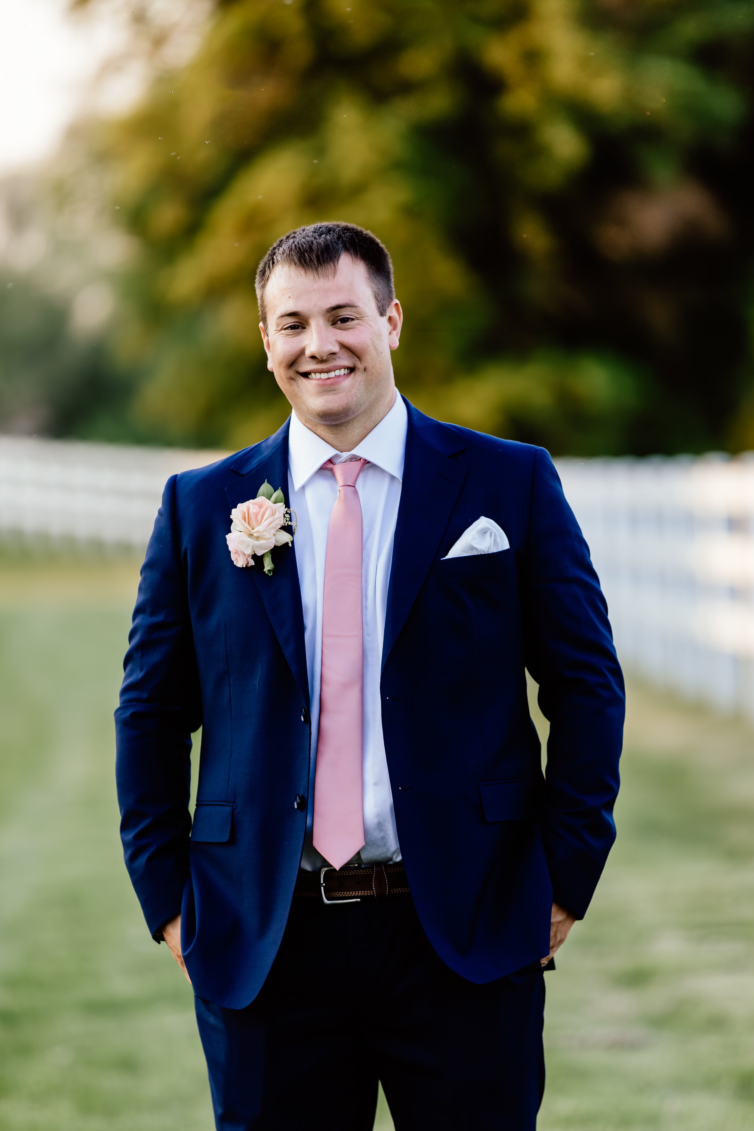 Smiling man in a dark blue suit, pink tie, with a white pocket square and pink boutonniere, outdoors with green trees and a fence in the background at Laurel Ridge in Sioux Falls SD.