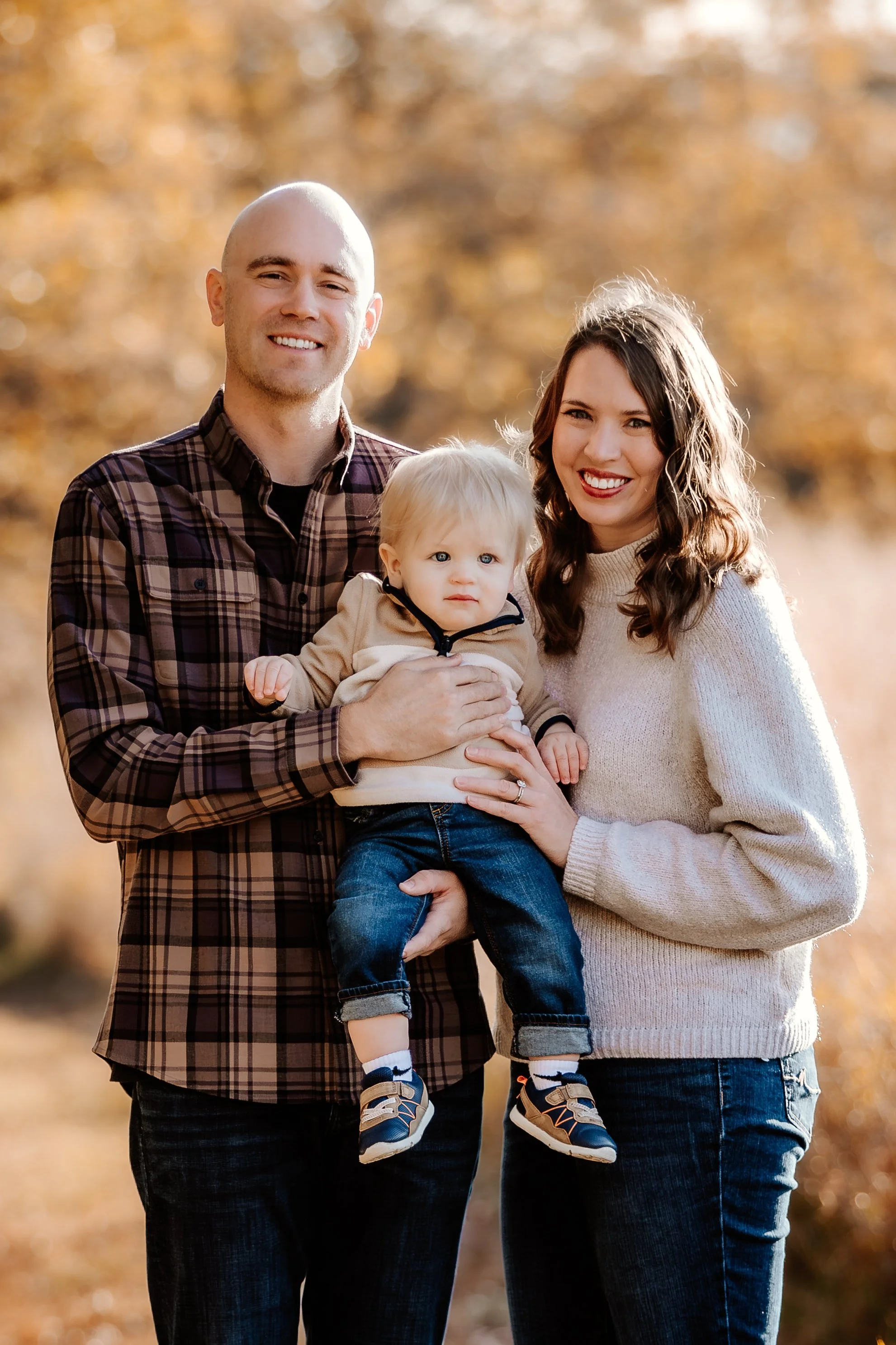 A family of three standing outdoors during fall, with golden autumn leaves in the background in Sioux Falls, South Dakota at Good Earth State Park.