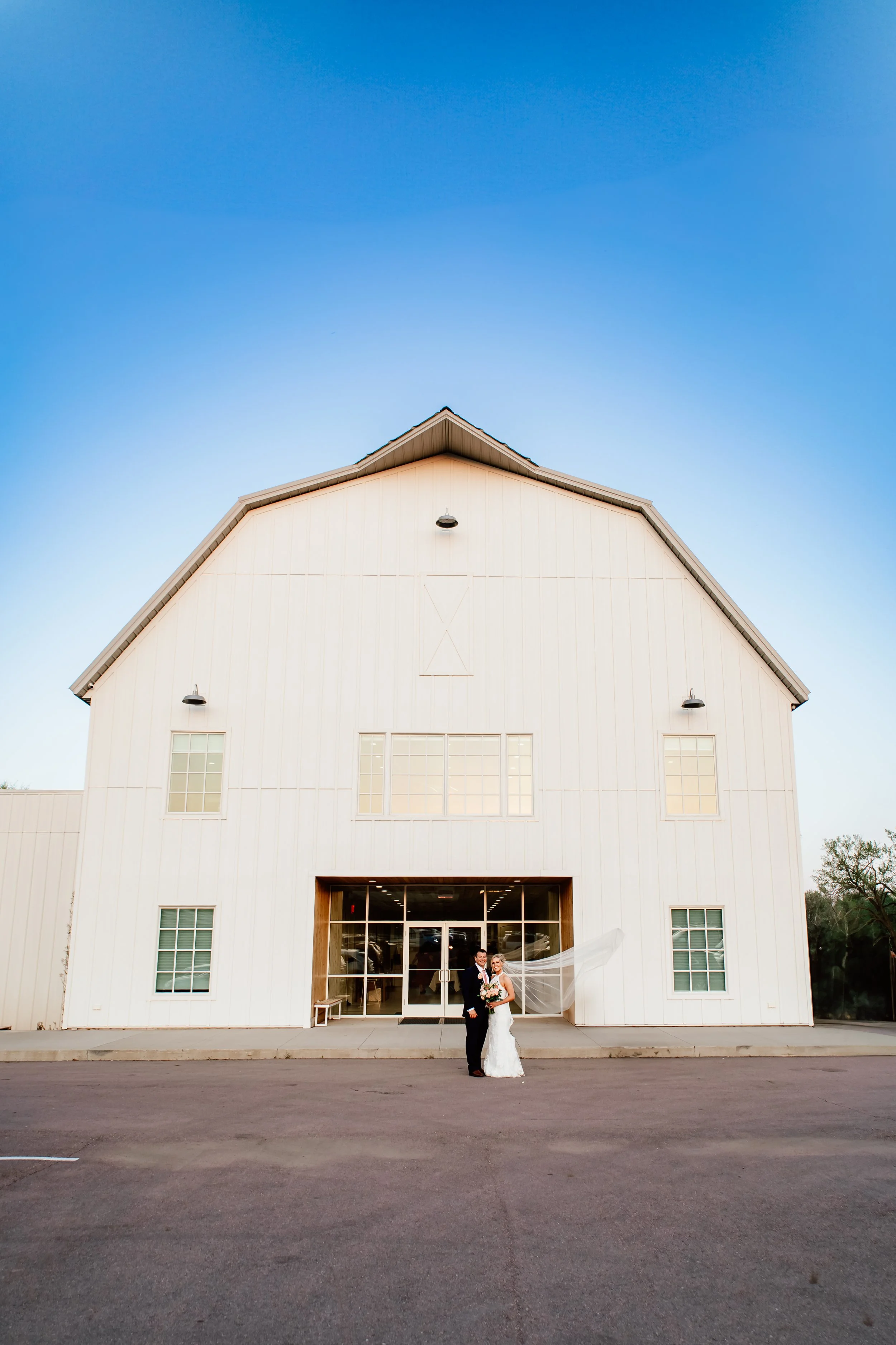 A bride and groom standing outside a large white barn, with the bride wearing a veil and holding a bouquet, smiling at the camera during their wedding photo in Sioux Falls, South Dakota at Laurel Ridge.