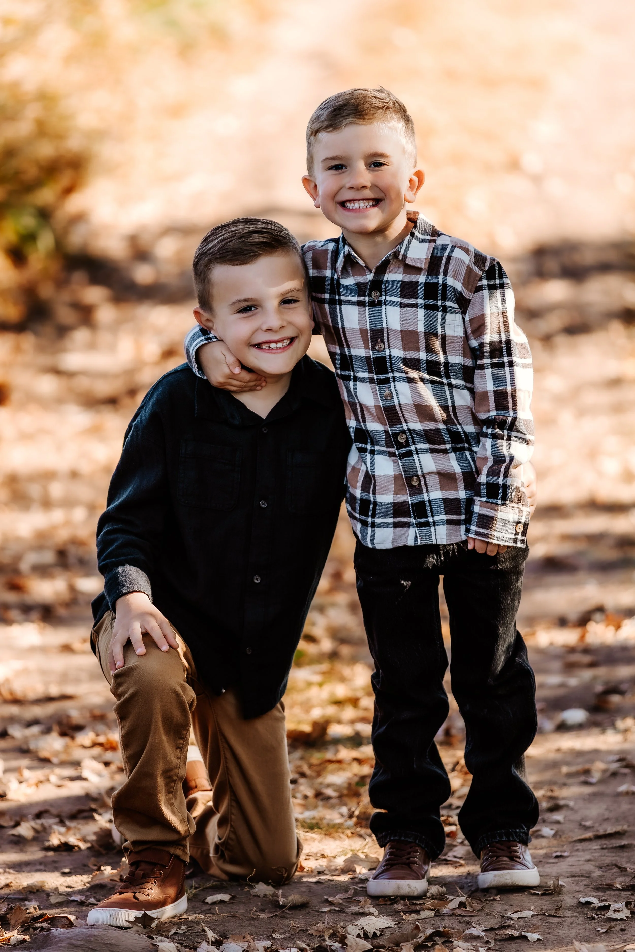 Two young boys smiling, one kneeling and the other standing, outdoors in a fall setting with fallen leaves in Sioux Falls, South Dakota at Good Earth State Park.