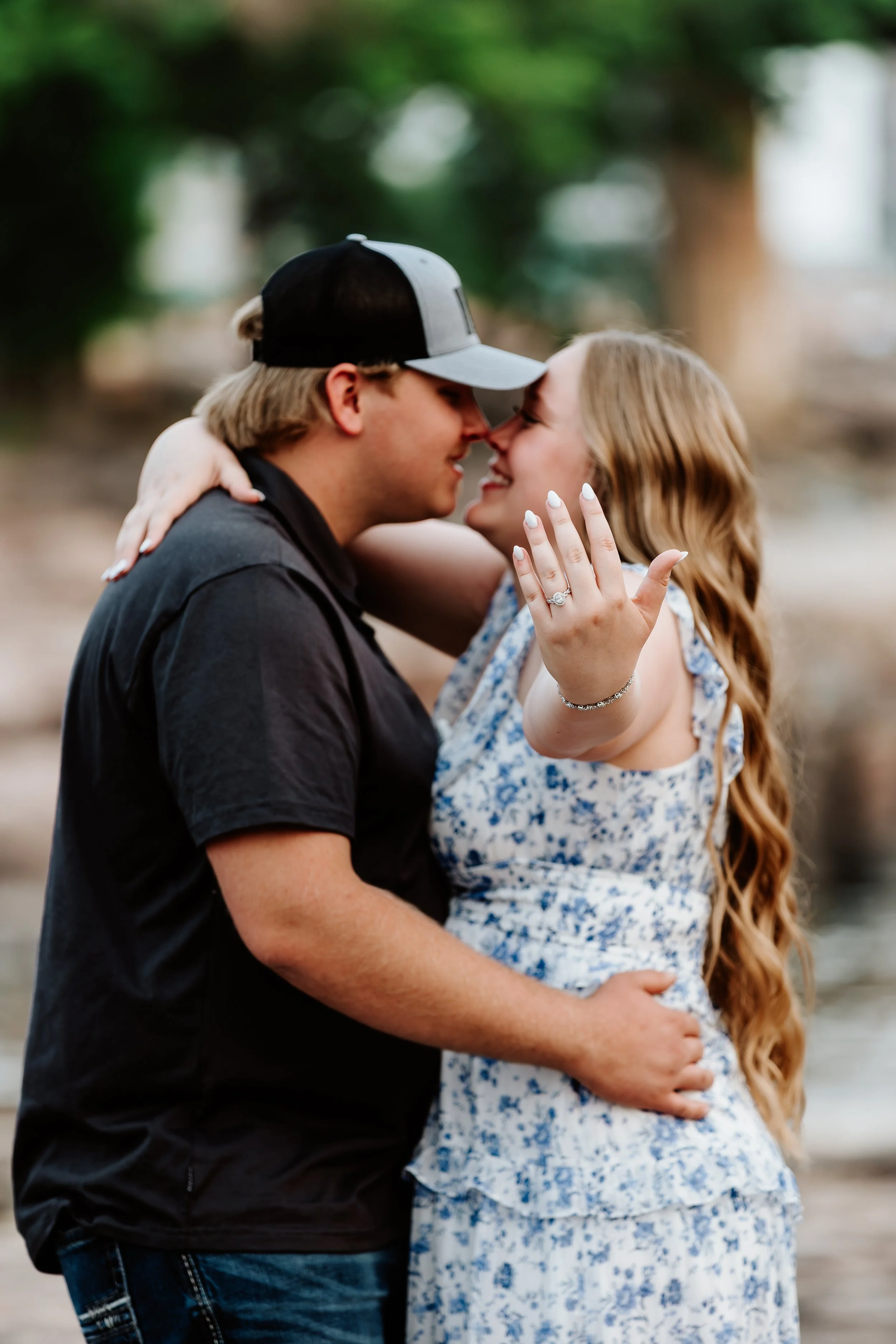 A couple embracing outdoors, smiling, with the woman showing off an engagement ring at The Falls Park in Sioux Falls, South Dakota.