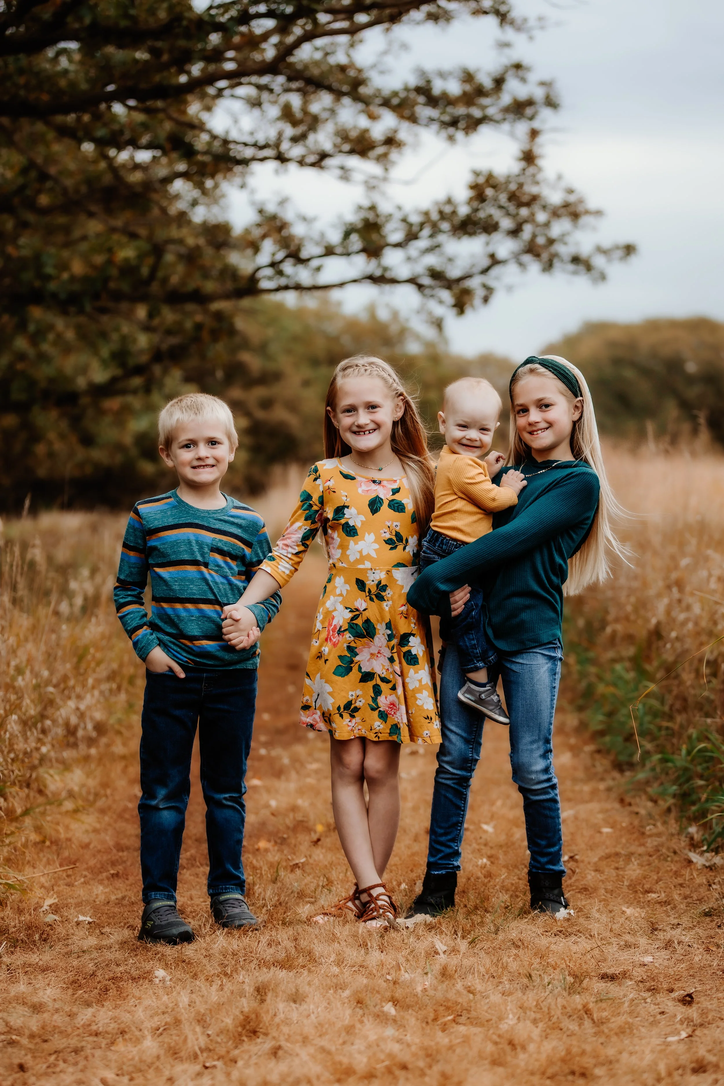 Four children standing on a dirt path in a park, holding hands and smiling, surrounded by trees with autumn foliage in Sioux Falls, South Dakota at Good Earth State Park.