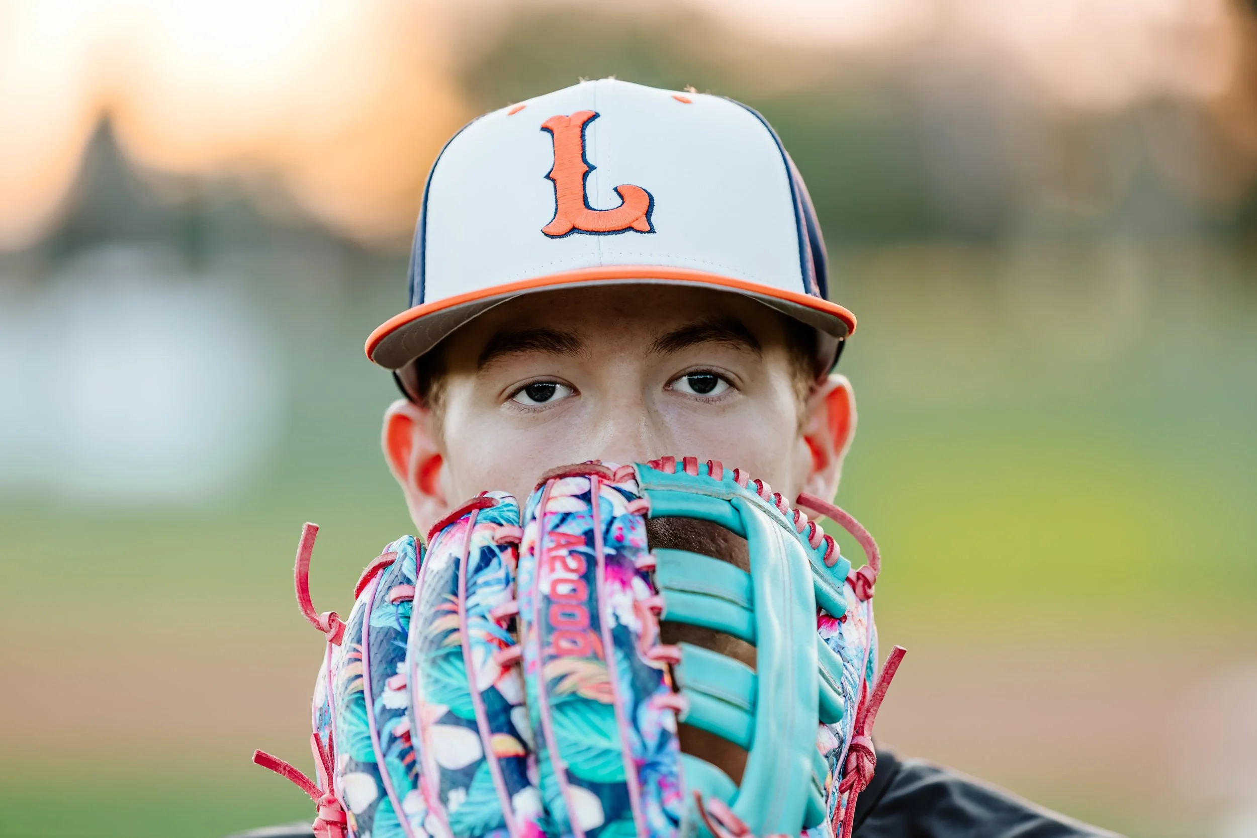 A young baseball player wearing a gray cap with an orange letter 'L' on it, holding a colorful baseball glove in front of their face, outdoors during sunset in Lennox, South Dakota.