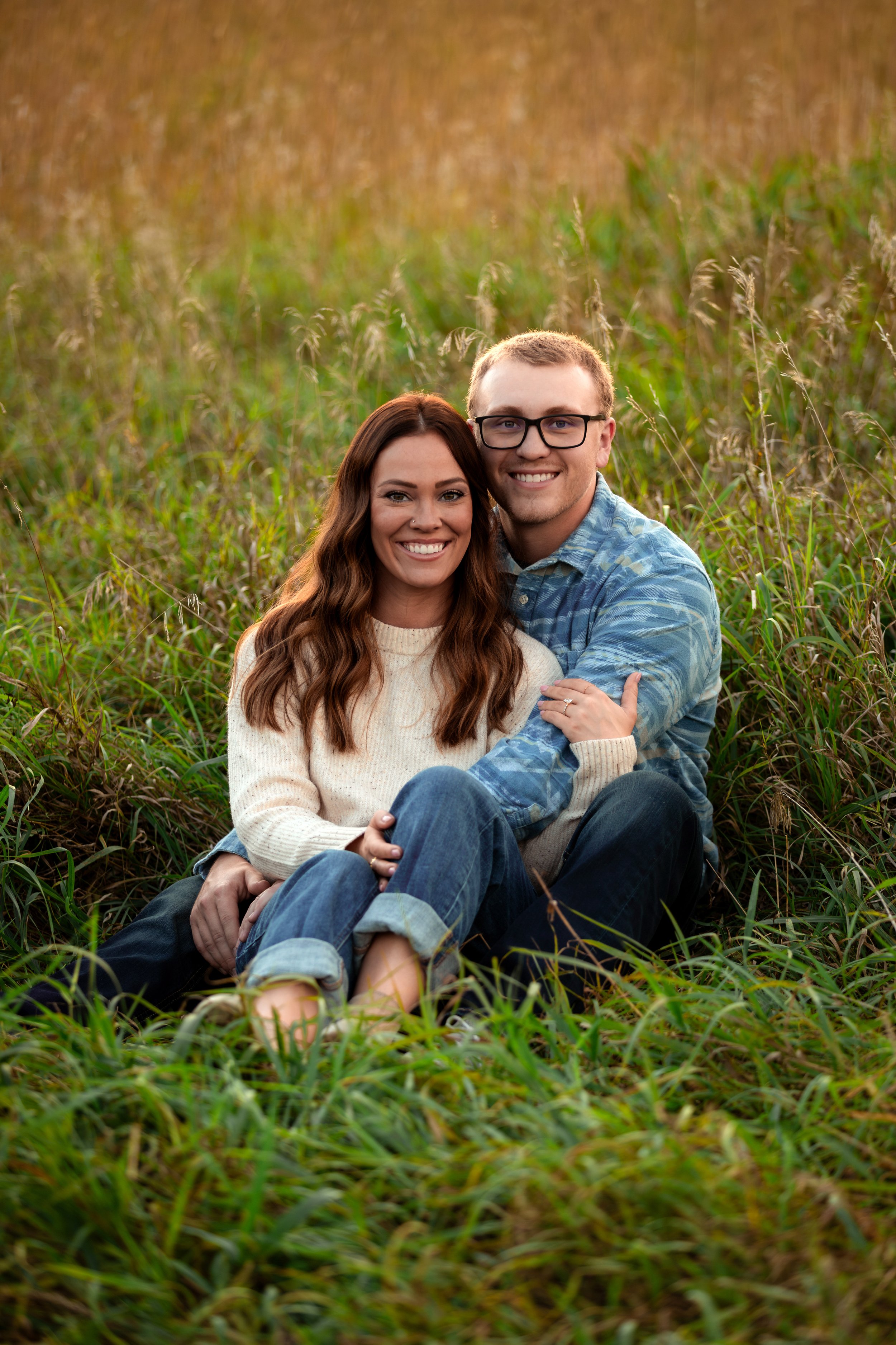 A smiling couple sitting in a grassy field during sunset, embracing each other at Good Earth State Park in Sioux Falls, South Dakota.