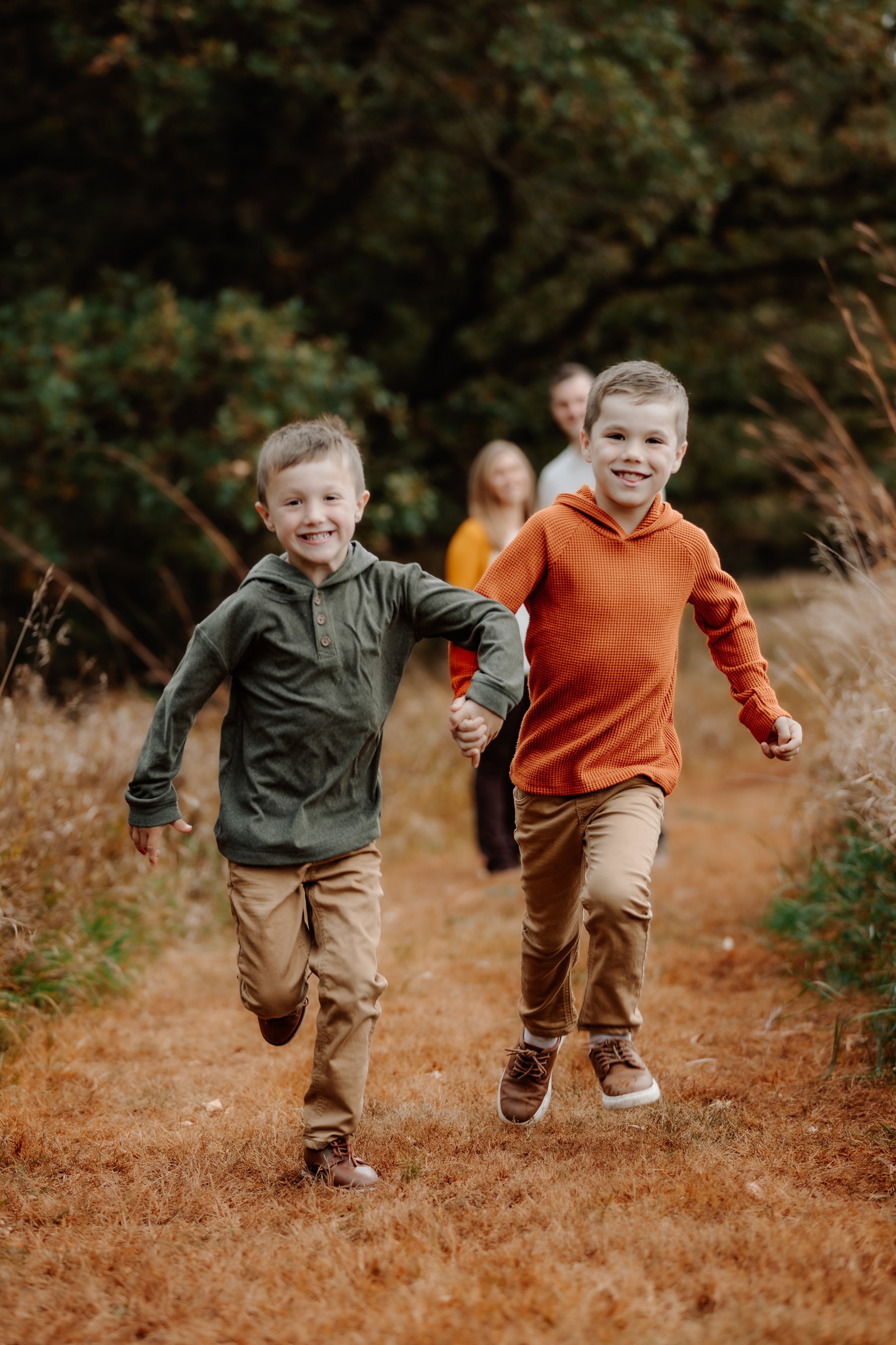 Two young boys run happily through a wooded path holding hands, with two adults in the background during an autumn day in Sioux Falls, South Dakota at Good Earth State Park.
