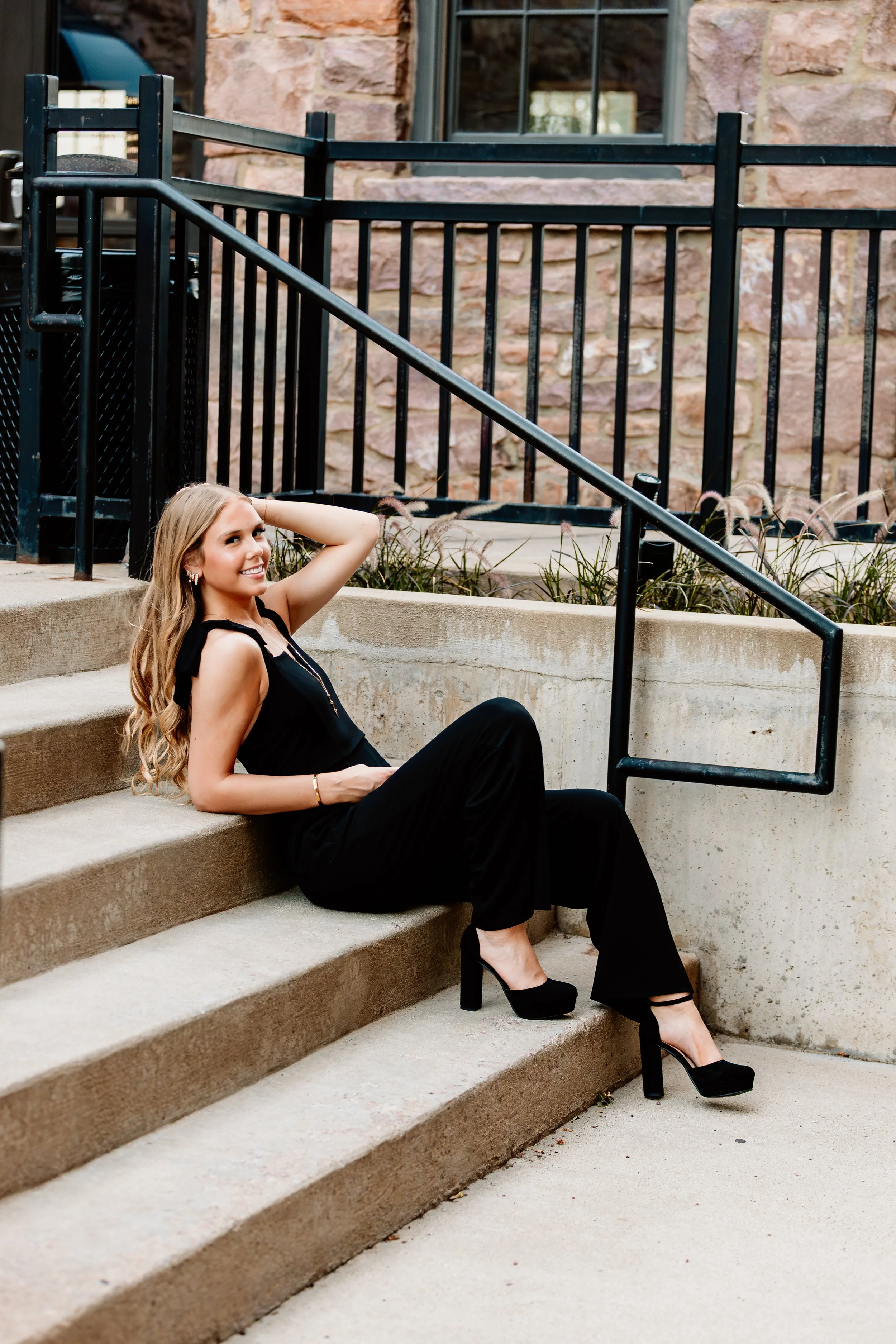 A woman with long blond hair in a black outfit and high heels is sitting on outdoor stairs, smiling, with a black railing and a stone building in the background in Downtown Sioux Falls, South Dakota.
