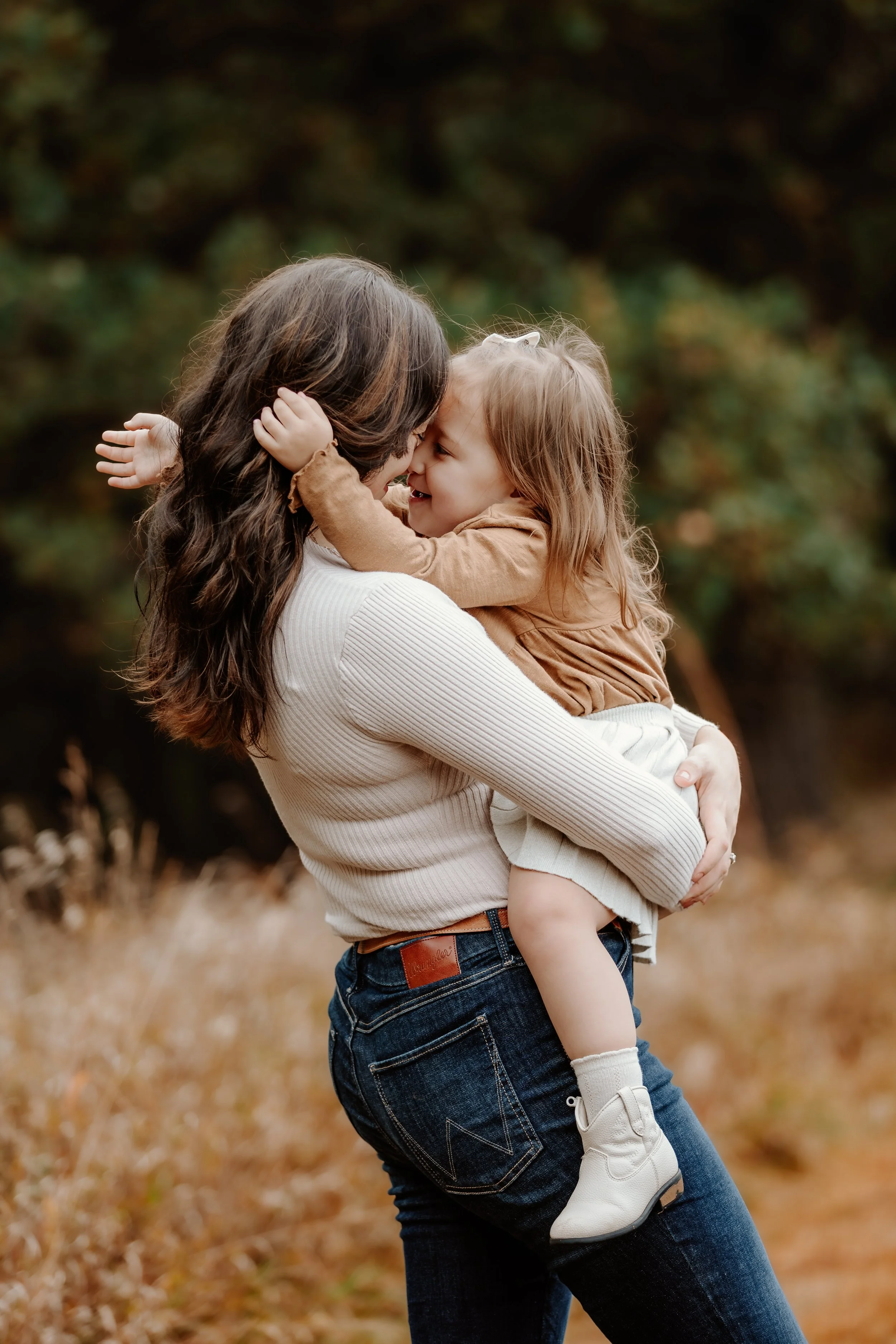 A woman holding a young girl outdoors during autumn, both smiling and touching noses, with trees and fall foliage in the background in Sioux Falls, South Dakota at Good Earth State Park.