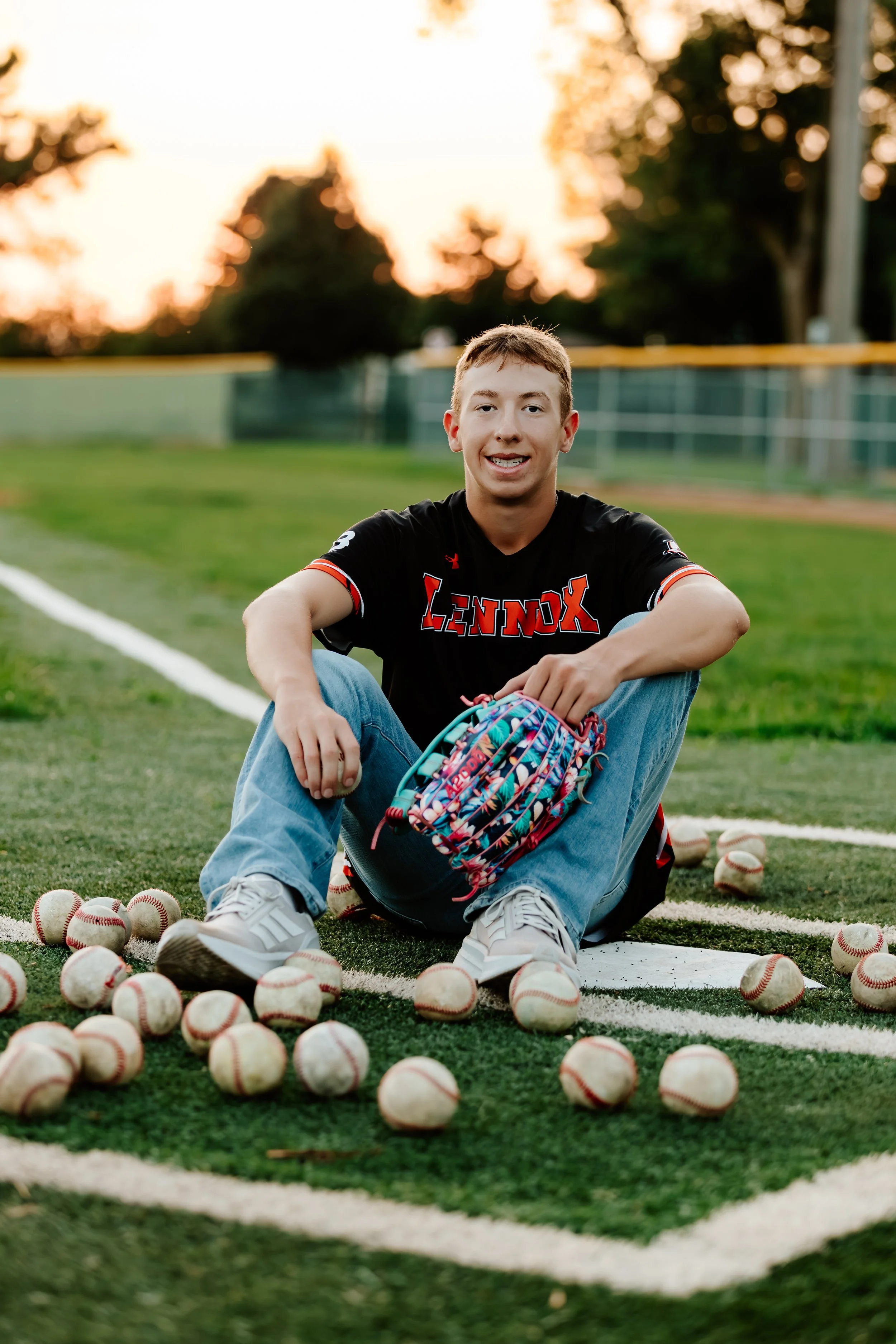 A young male baseball player sitting on a baseball field at sunset, surrounded by baseballs and holding a colorful baseball glove, wearing a black and red team jersey in Lennox, South Dakota.