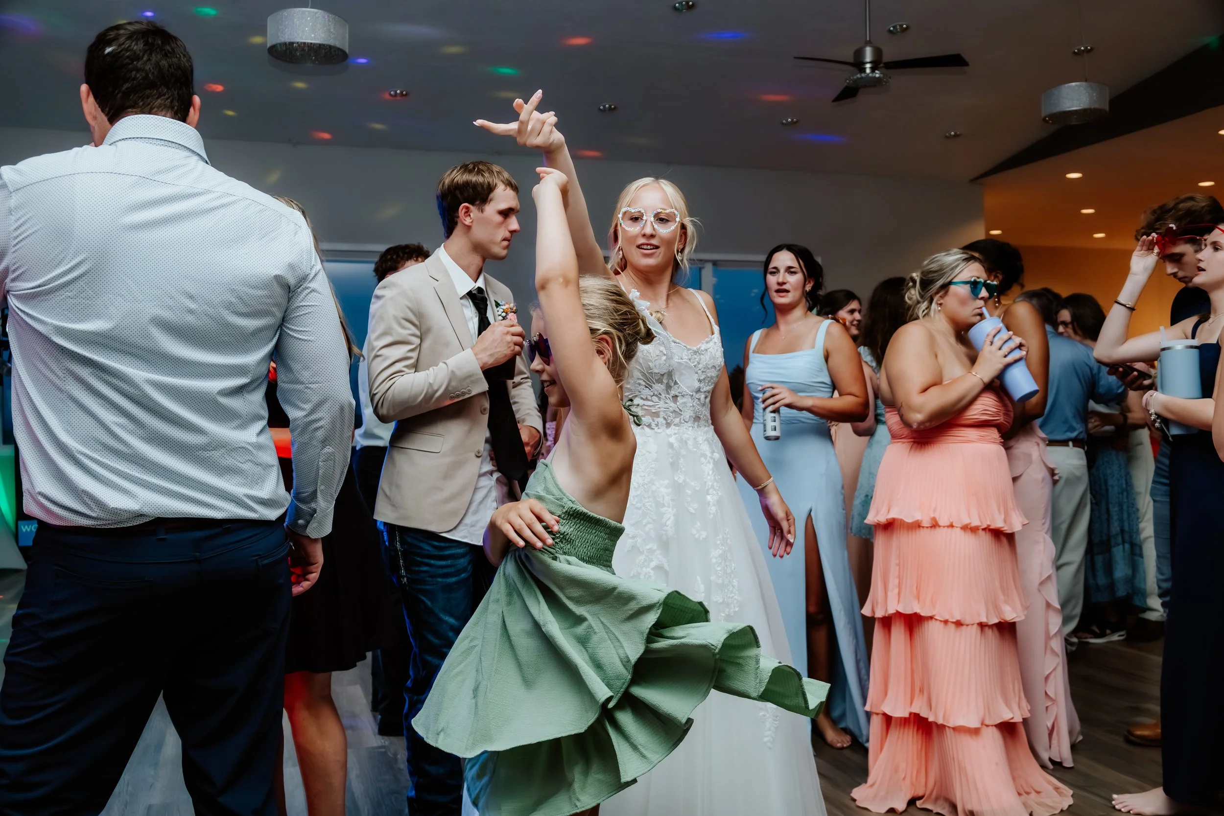 People dancing and socializing at a party, including a woman in a white dress, a girl in a green dress dancing, and others holding drinks in a decorated indoor venue in Luverne, MN.