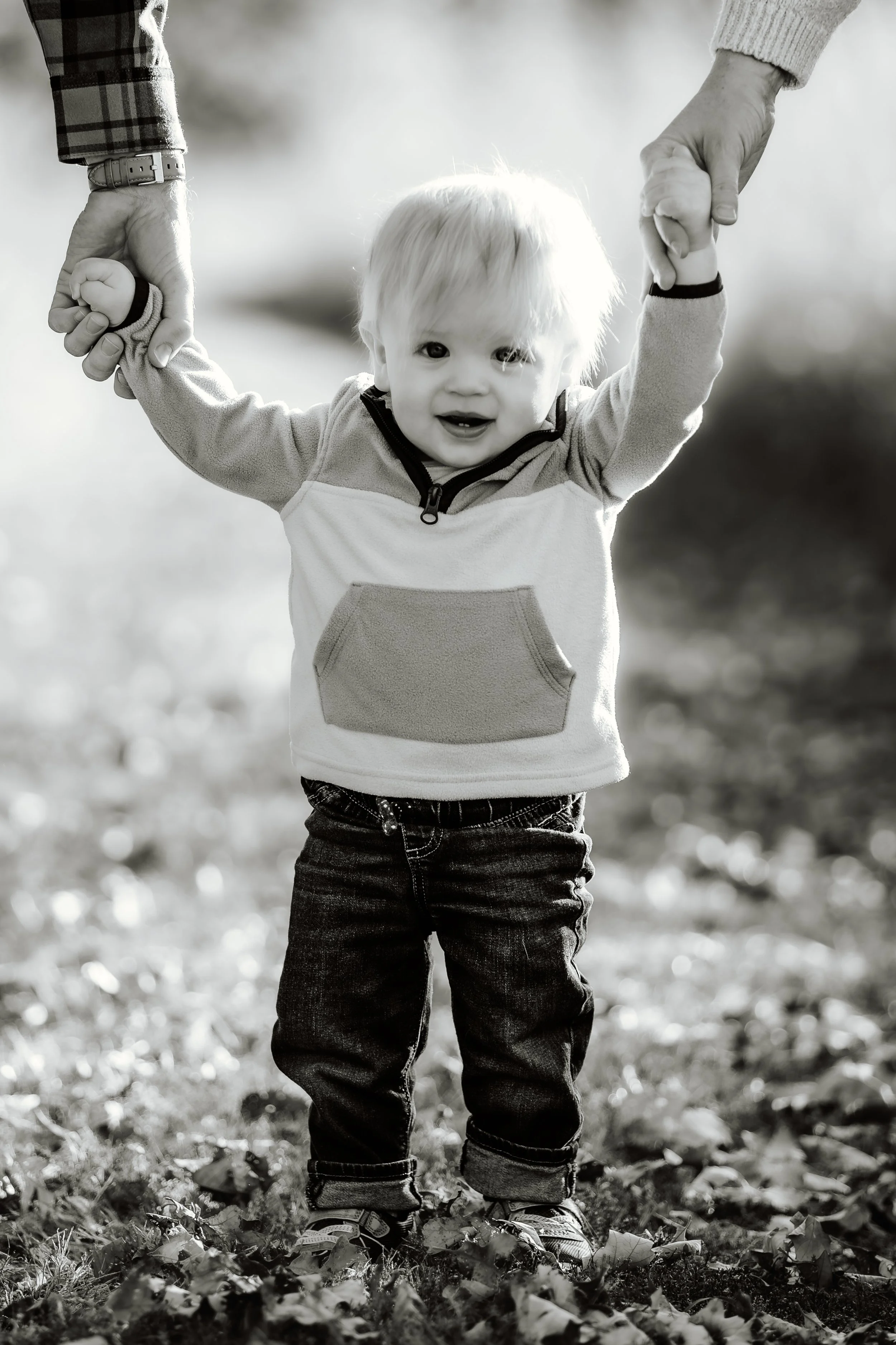 A young child with blond hair standing outdoors on a leaf-covered ground, holding hands with two adults, smiling happily in Sioux Falls, South Dakota at Good Earth State Park.