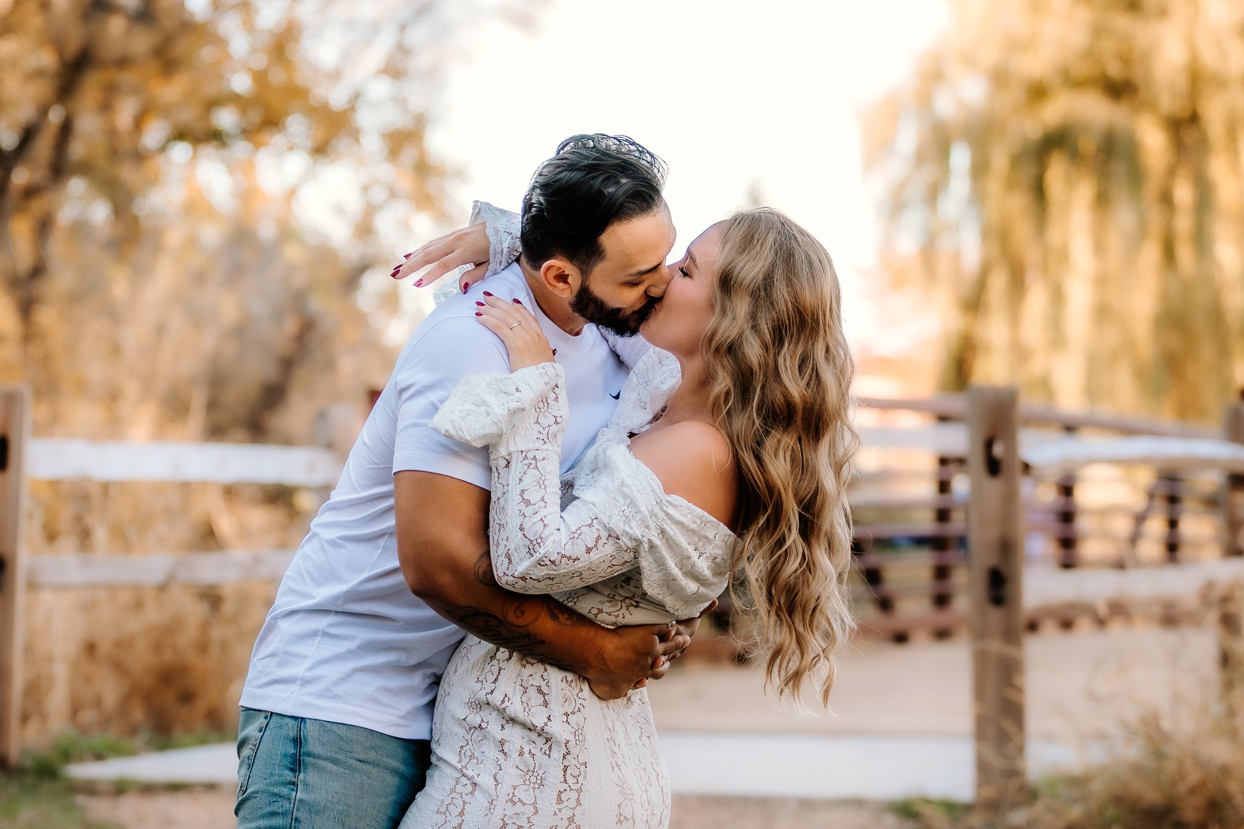 A couple sharing a kiss outdoors during sunset, with autumn trees and a wooden fence in the background at Mary Jo Wegner Arboretum in Sioux Falls, South Dakota.