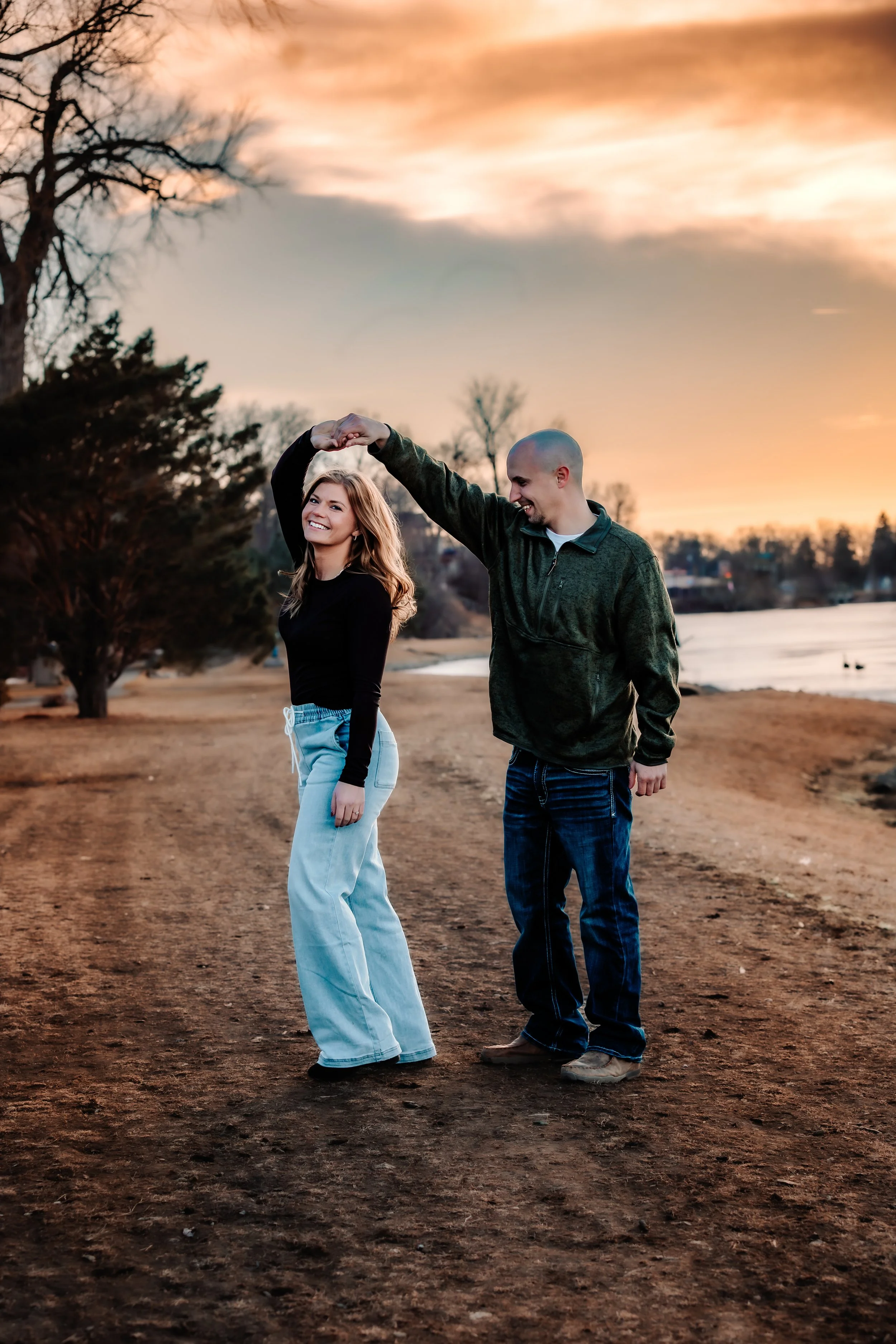 A young couple dancing and smiling on a dirt path near a lake during sunset at Terrace Park in Sioux Falls, South Dakota.